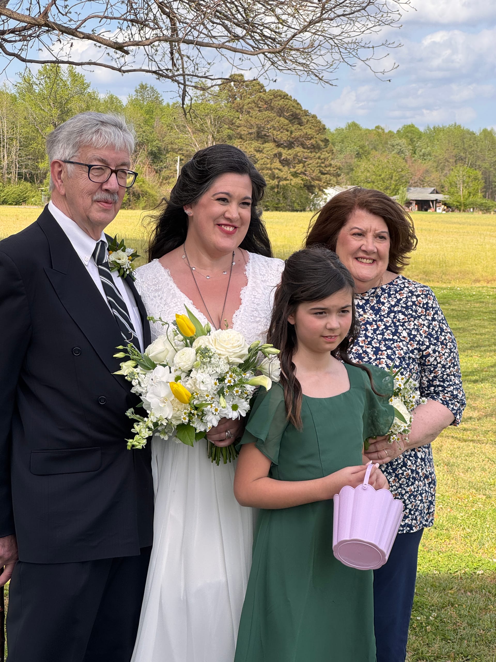 Bride in white lace gown holding a yellow and white bouquet poses with family including a flower girl in green dress against a sunny farmland field backdrop at Scarborough House.