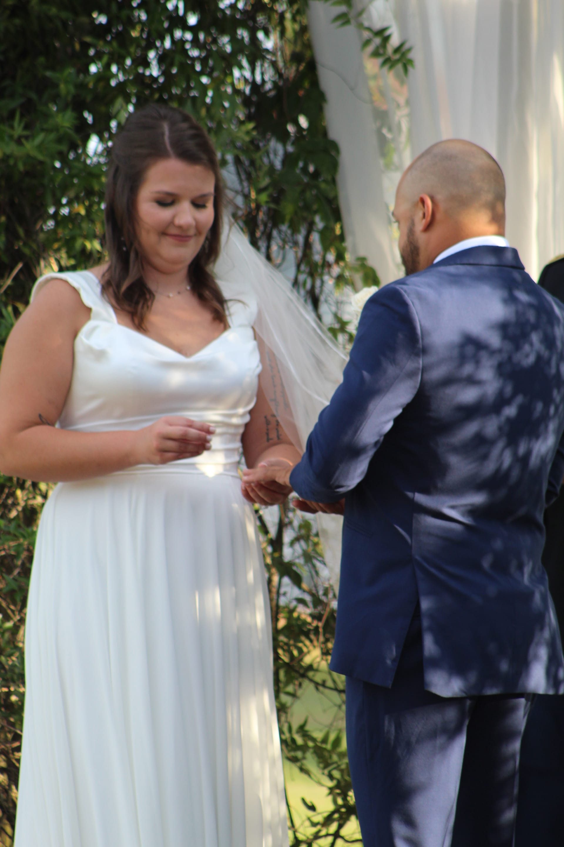 Bride in a flowing white gown and veil smiles softly as groom in a navy suit places a ring on her finger during an outdoor wedding ceremony at Scarborough House.
