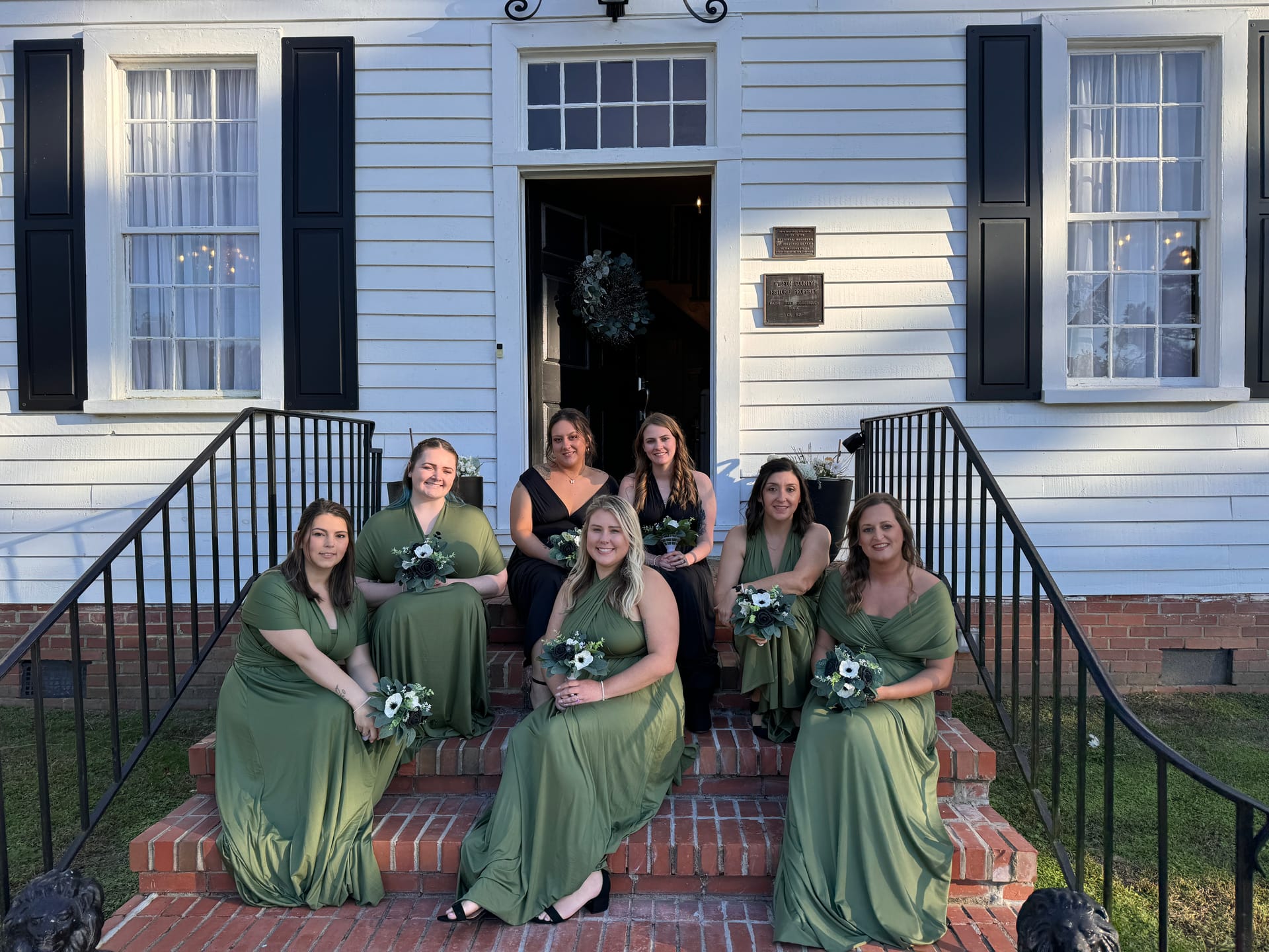 Seven bridesmaids in sage green wrap gowns holding white anemone bouquets posed on the brick front steps of the 1821 Scarborough House manor.