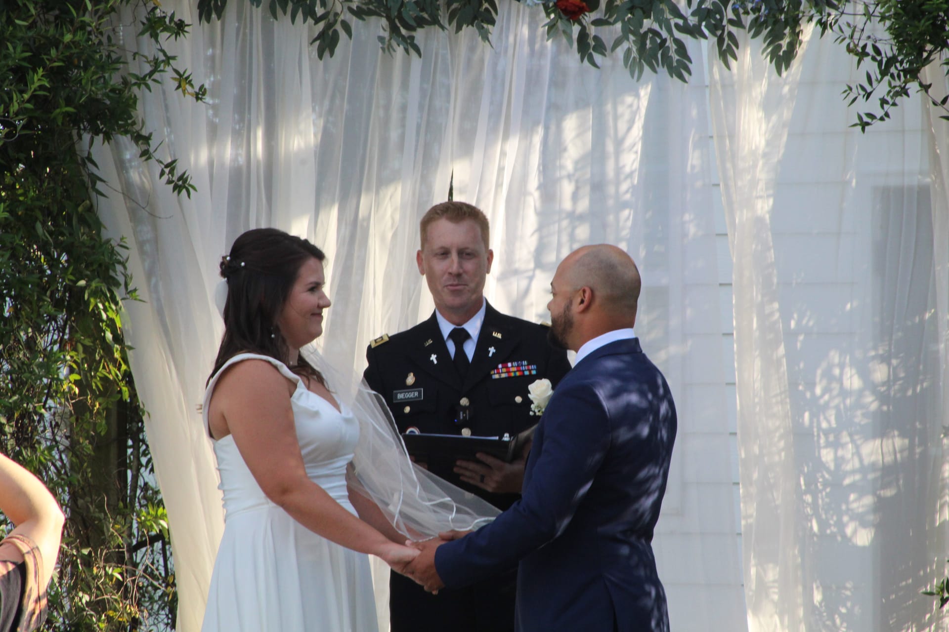 Bride and groom hold hands and exchange vows before a military chaplain officiant beneath a white draped arch adorned with greenery at Scarborough House.