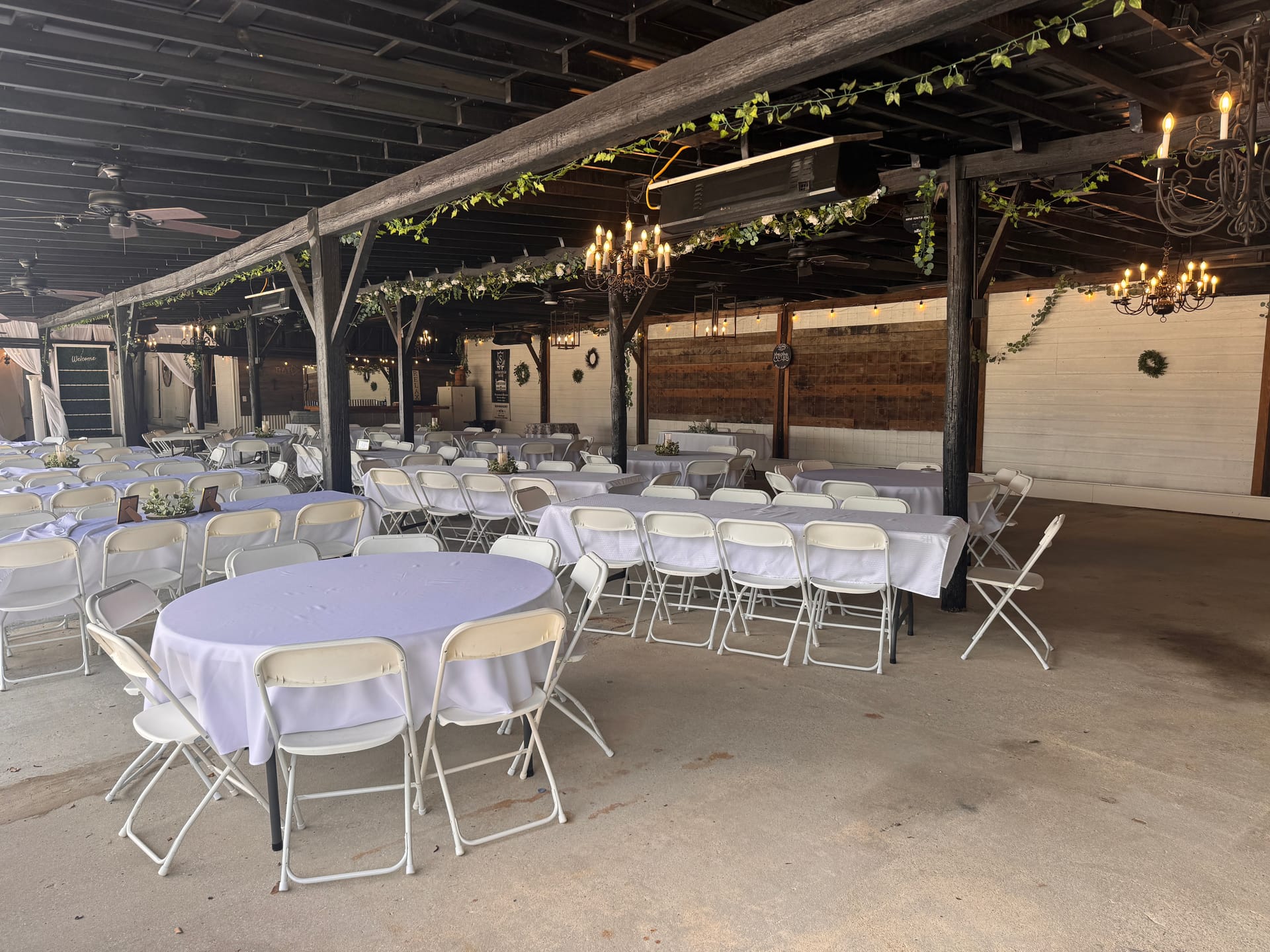 Rustic-elegant magnolia pavilion at Scarborough House set for a wedding reception with white linen round and banquet tables, folding chairs, greenery-draped beams, and wrought-iron candle chandeliers.