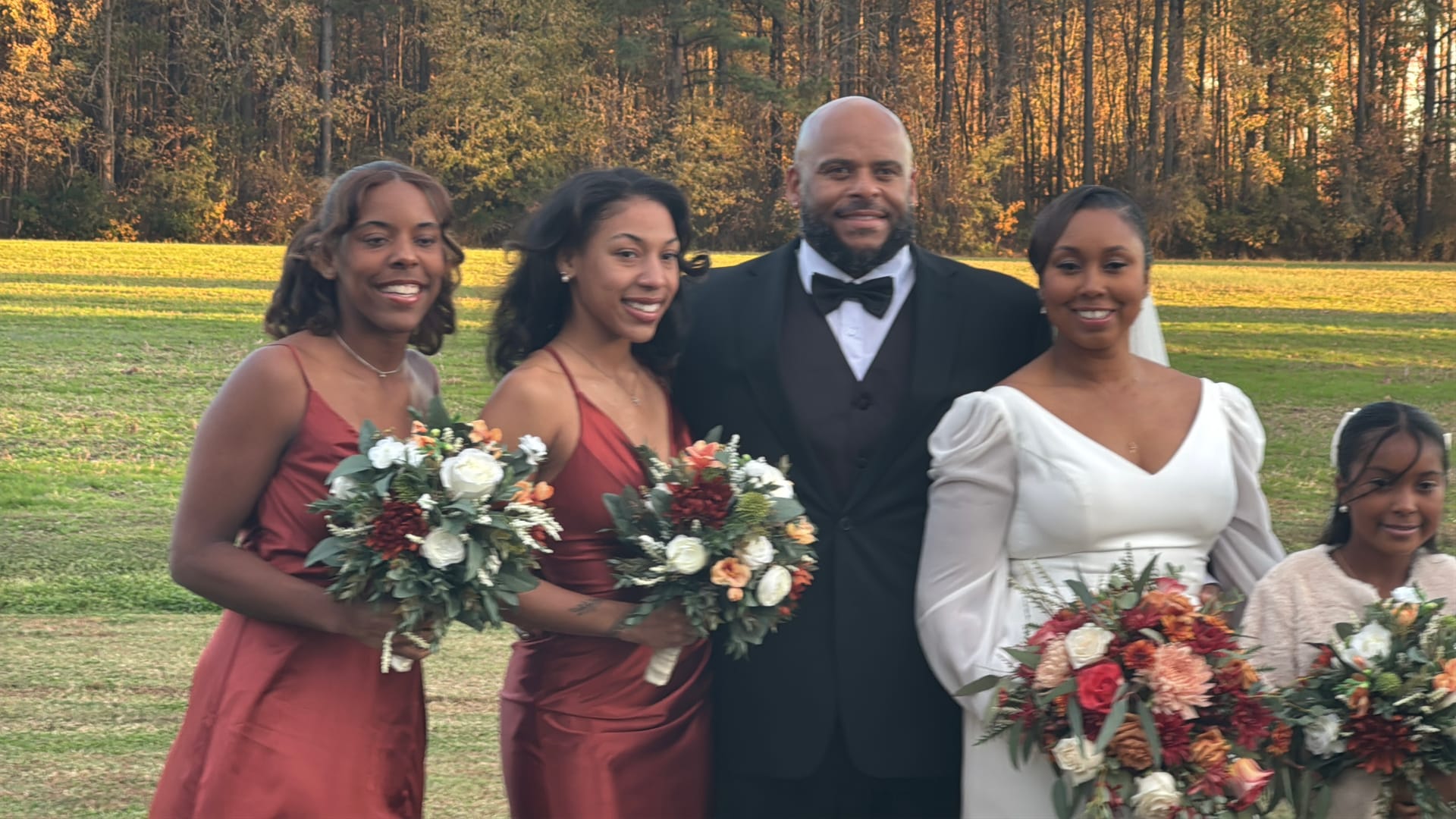 Bride in white gown with groom in black tuxedo and two bridesmaids in rust-colored dresses holding floral bouquets posing on the ceremony lawn at Scarborough House during golden hour with autumn trees in the background.