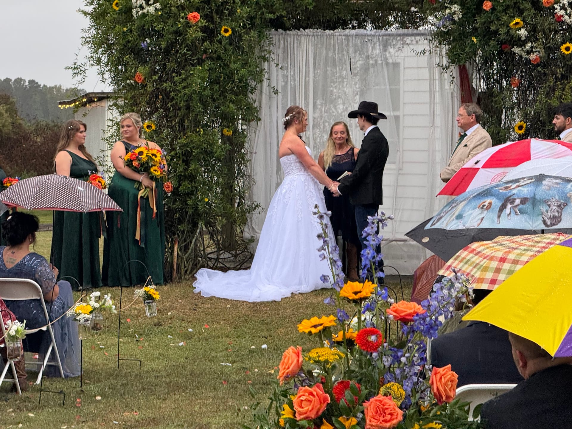 Bride and groom exchange vows beneath a lush floral arch adorned with sunflowers and greenery on the ceremony lawn at Scarborough House as bridesmaids in emerald green look on.