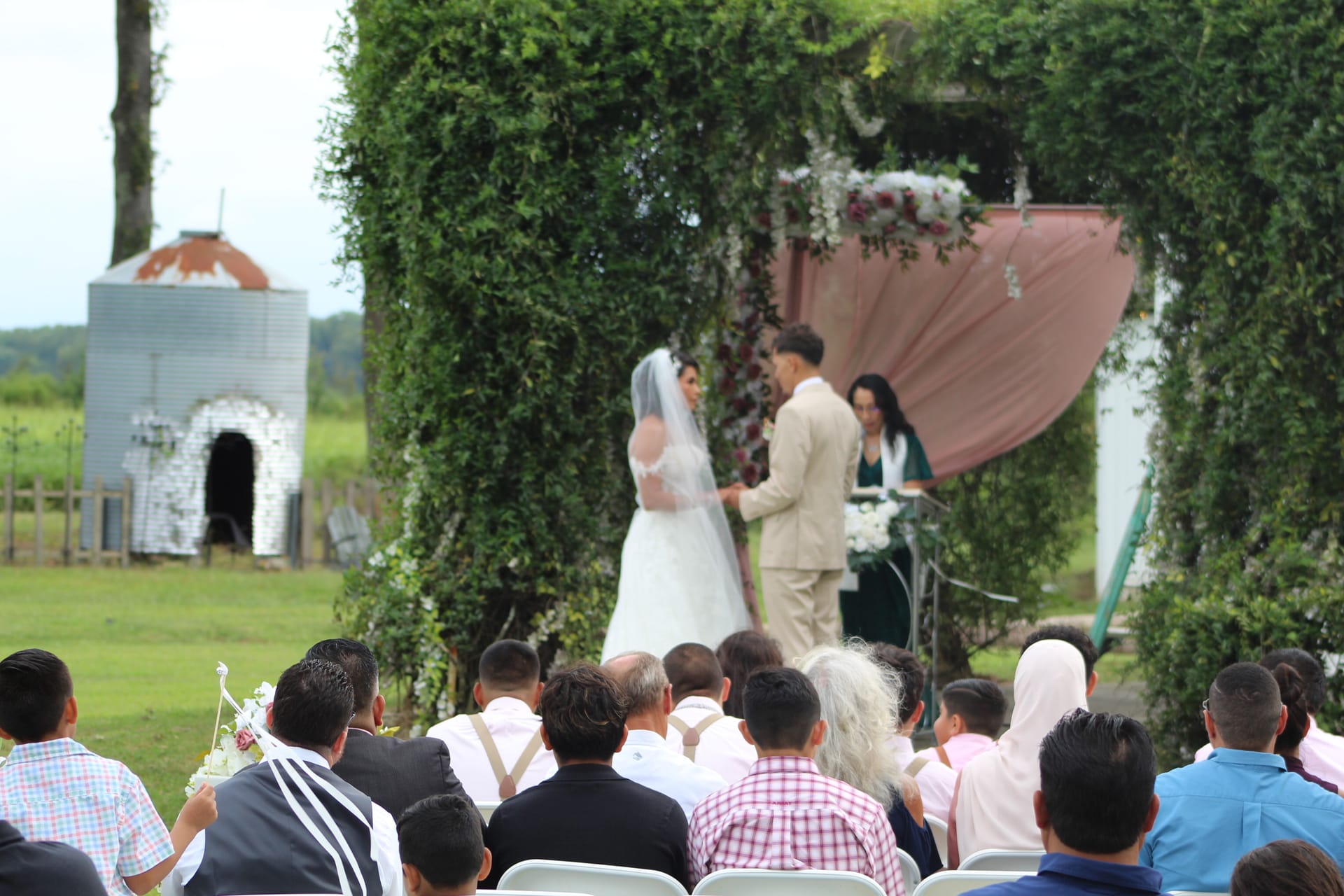 Bride and groom exchanging vows beneath a lush ivy-covered arch adorned with florals and blush drapery on the ceremony lawn at Scarborough House, surrounded by seated guests.