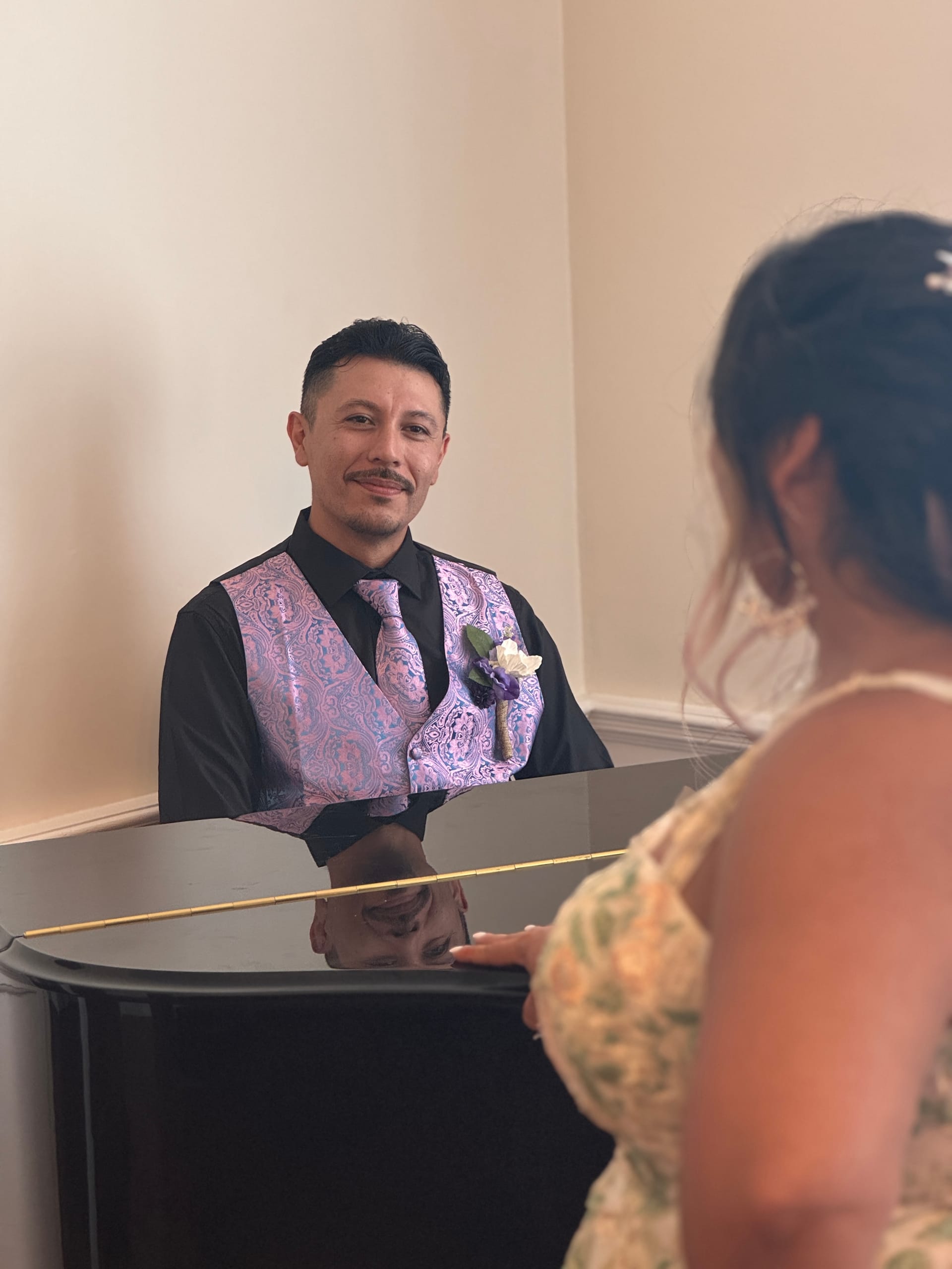 Groom in a purple paisley vest and black shirt smiles warmly across a grand piano toward his bride during an intimate first-look moment inside the historic manor.