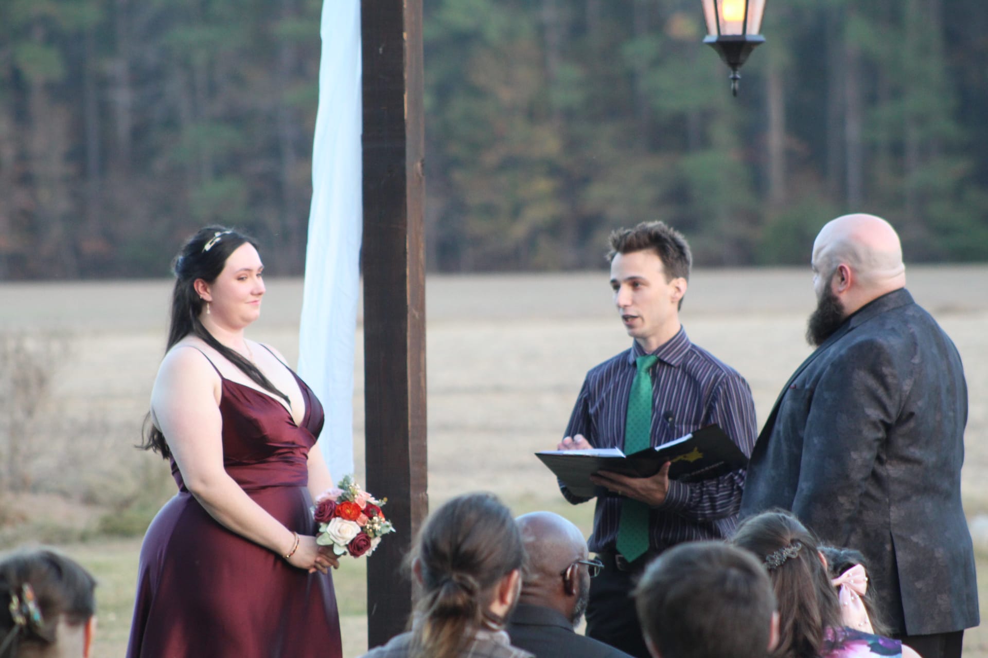 Bride in a burgundy gown holding a red and white bouquet stands at an outdoor evening ceremony while an officiant reads from a book and the groom looks on, with wooded farmland fields in the background.