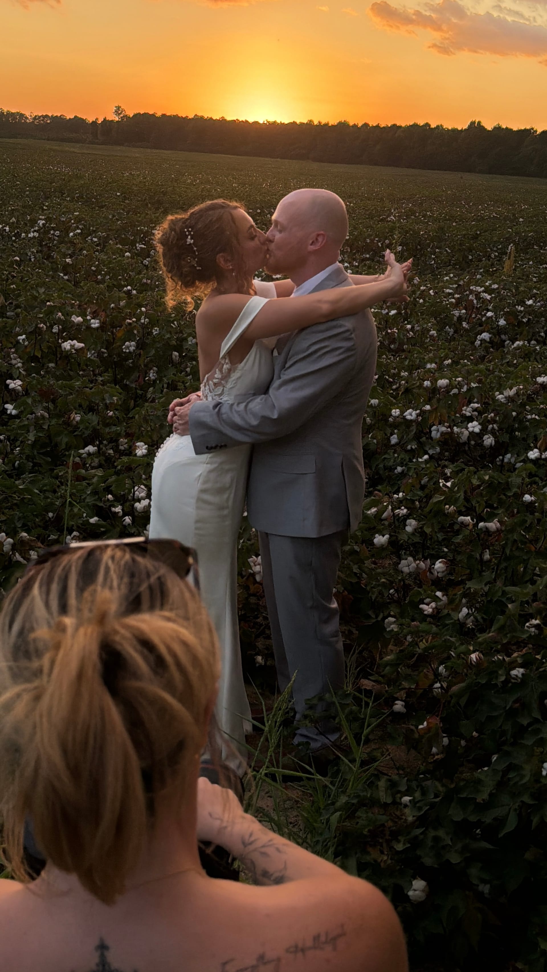 Bride and groom share a romantic kiss in a cotton field at sunset, silhouetted against a glowing orange sky with a photographer capturing the moment in the foreground.