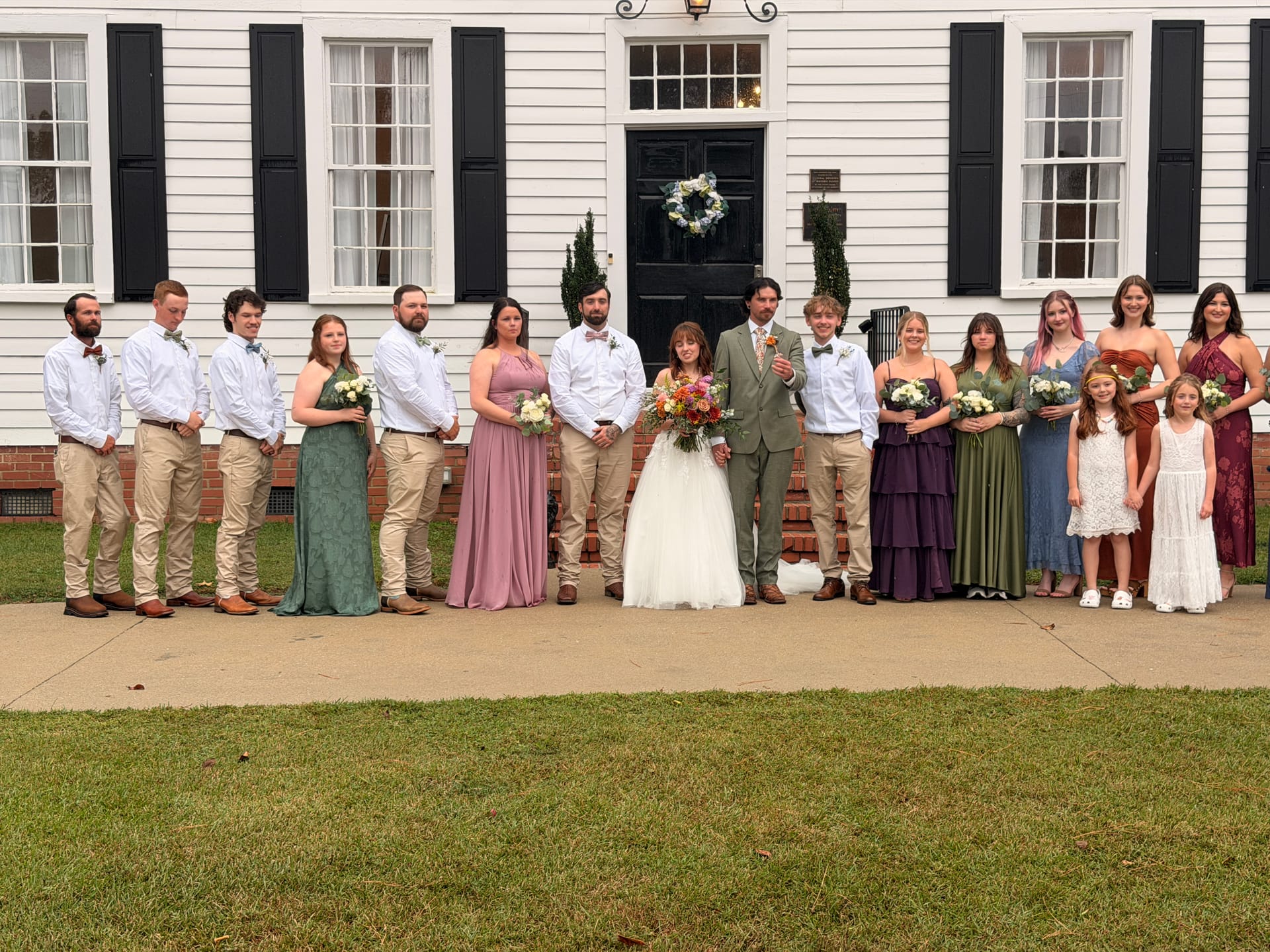 Full wedding party including bride and groom posed in front of the white 1821 Scarborough House manor with black shutters and a floral wreath on the door.