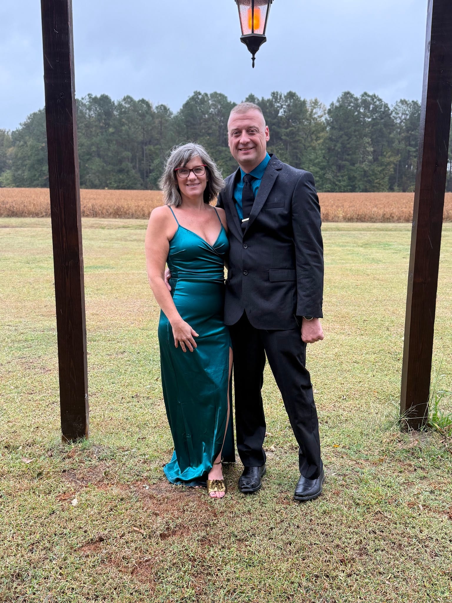 A couple dressed in a teal satin gown and charcoal suit pose together under a lantern structure with golden farmland fields and tree line behind them at Scarborough House.