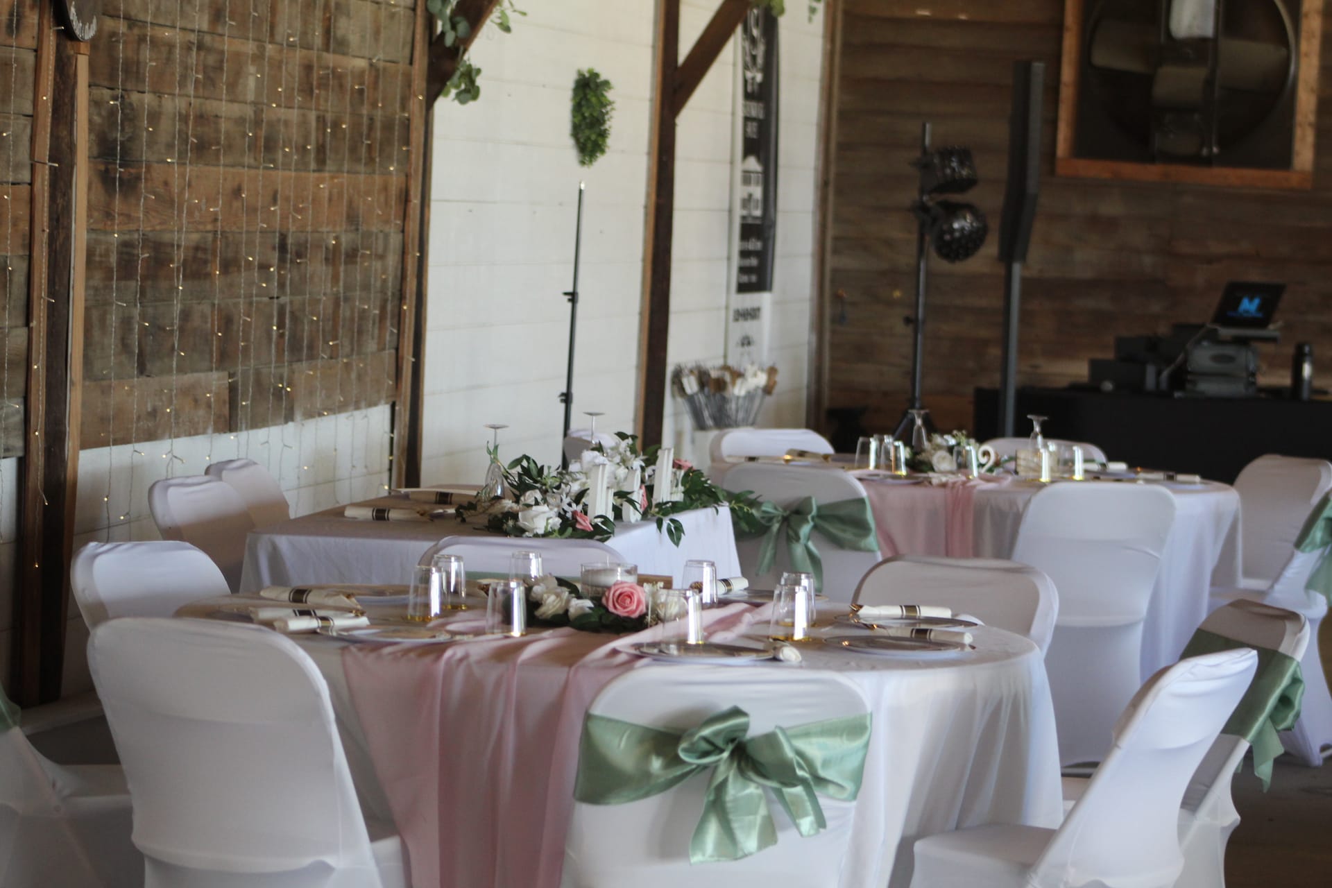 Elegantly styled wedding reception tables with white linens, sage green satin chair sashes, dusty rose table runners, floral centerpieces, and candles inside a rustic barn venue adorned with fairy lights.