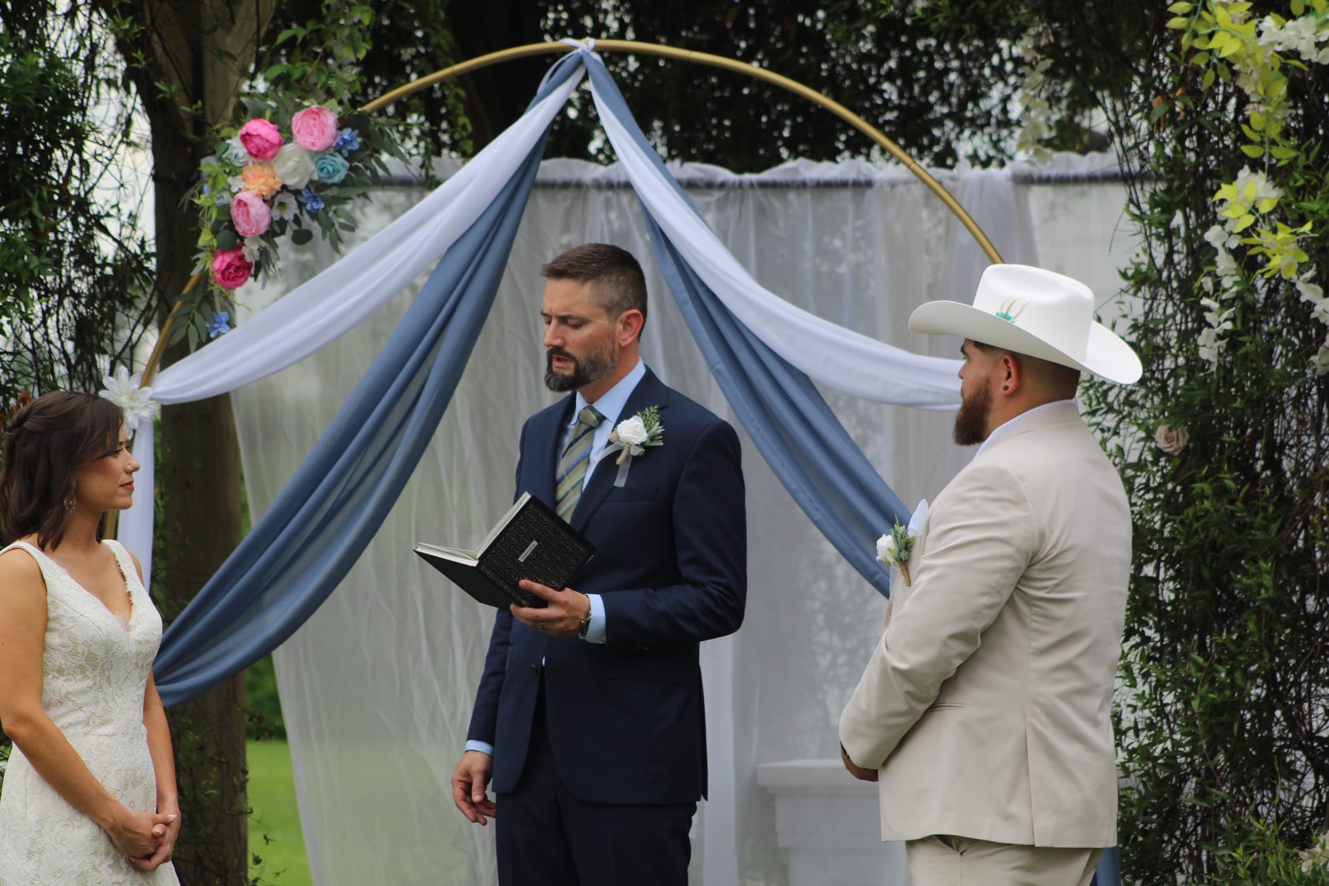 A bride in a white lace gown and groom in a tan suit and white cowboy hat stand before an officiant reading from a book beneath a gold circular arch draped in dusty blue fabric and colorful flowers at Scarborough House.