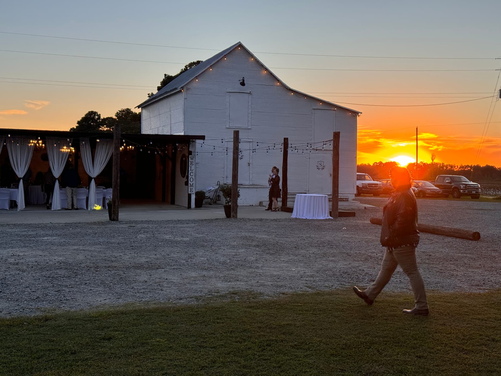 A silhouetted guest walks across a gravel path at sunset in front of a white barn wedding venue decorated with string lights, white draping, and chandeliers as the golden sun sets dramatically on the horizon.