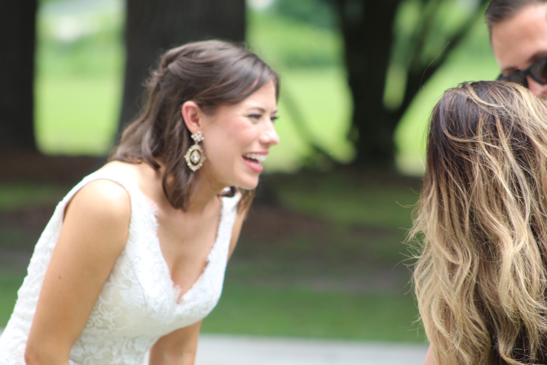 A radiant bride in a white lace gown laughing joyfully while leaning toward guests beneath the oak trees at Scarborough House estate.