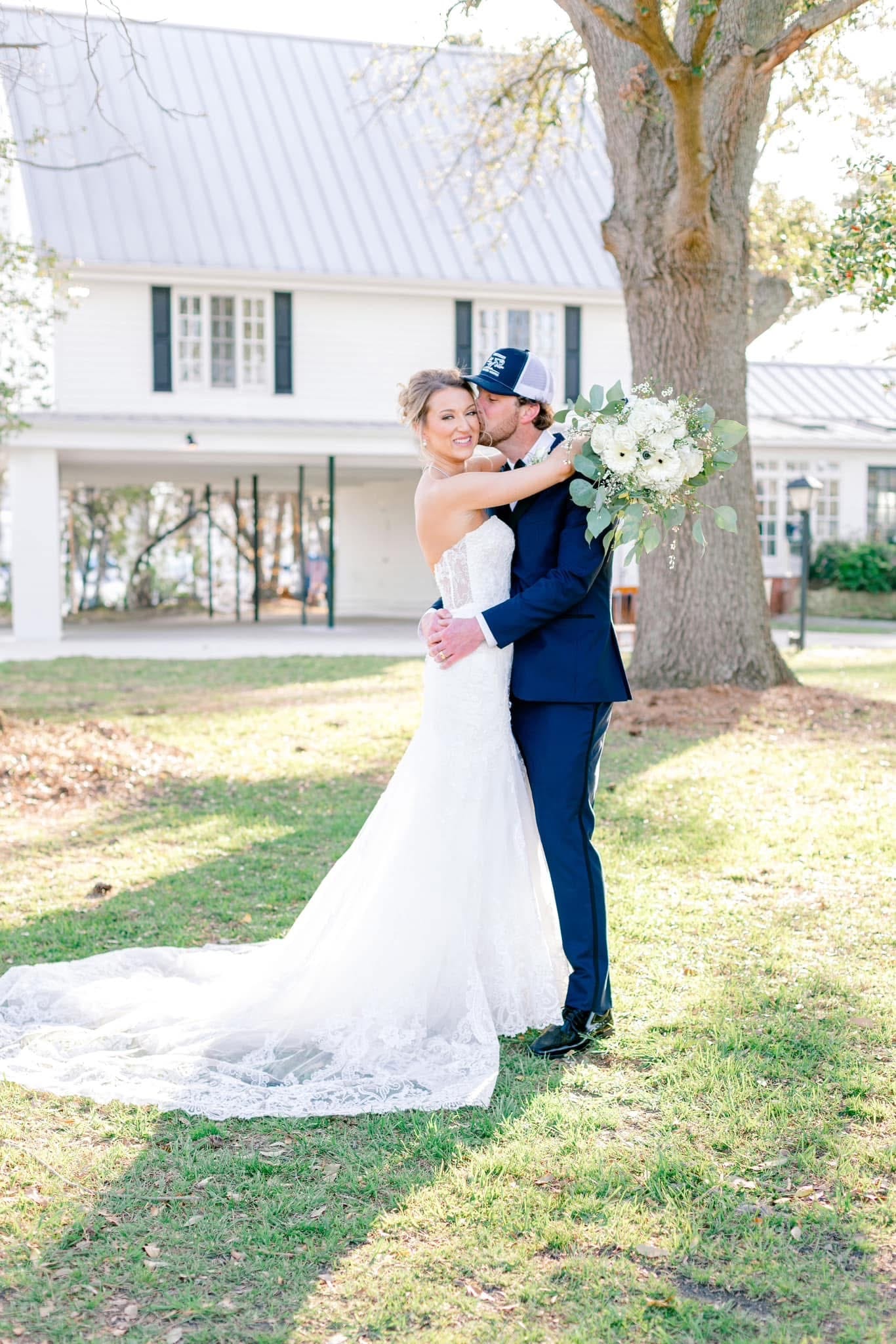 Bride in a strapless lace gown with cathedral train smiles as groom in a navy suit kisses her cheek beneath a grand oak tree with the white 1821 Scarborough House manor visible in the background.