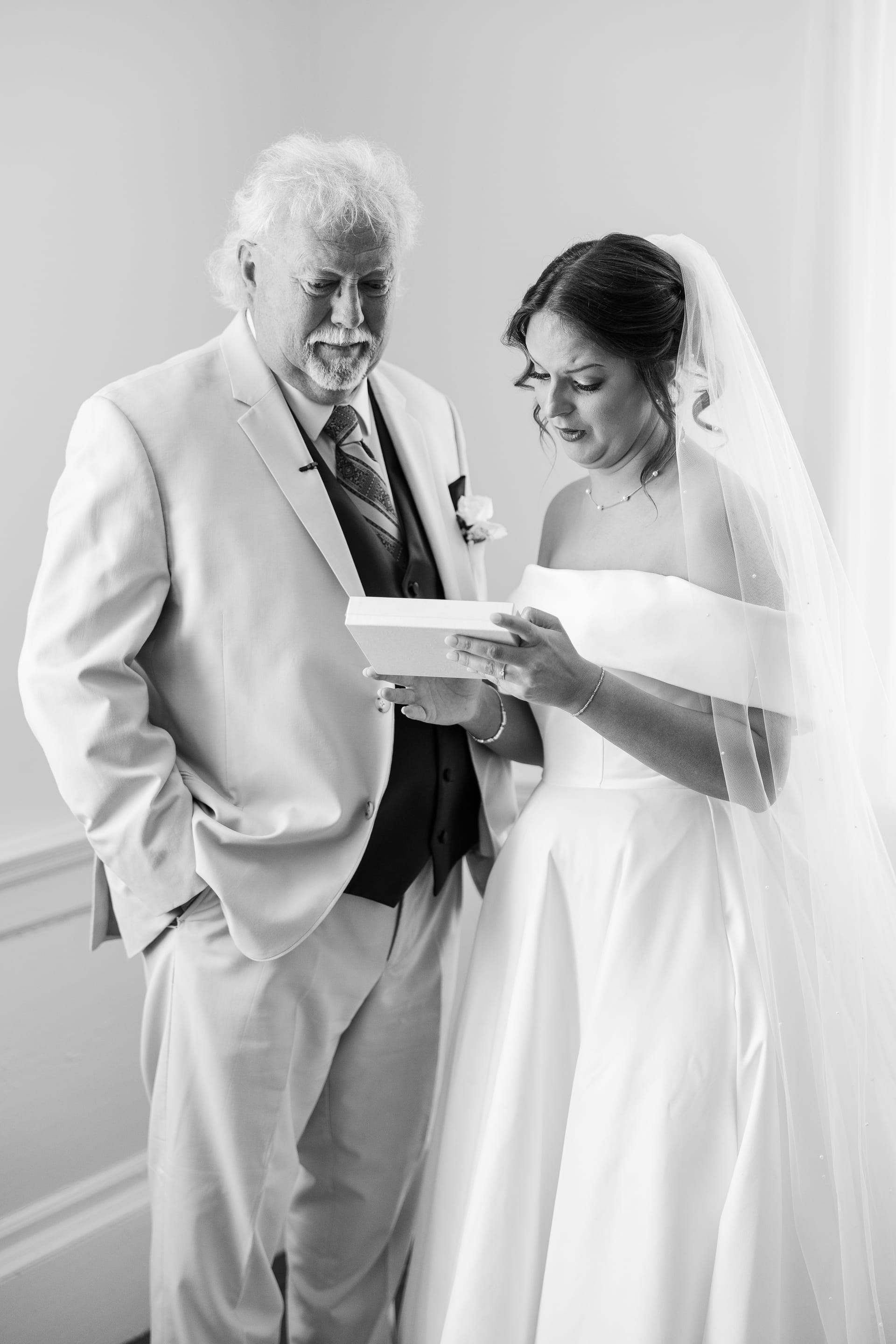 A bride in a white gown and veil reads a heartfelt note from a gift box alongside an older gentleman in a suit inside the bridal suite at Scarborough House.
