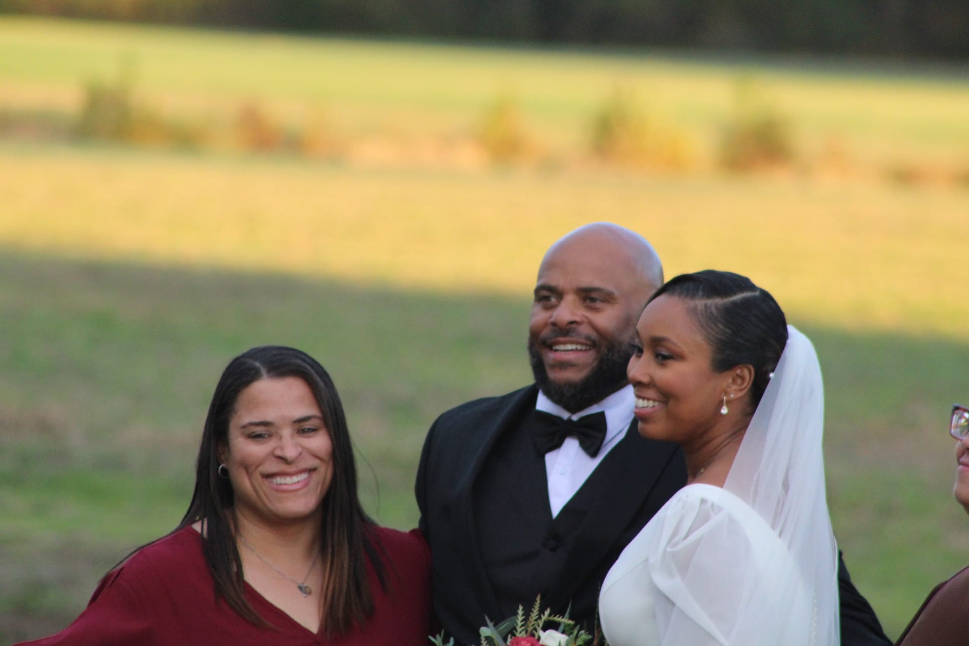 Smiling bride in white gown and veil and tuxedo-clad groom pose joyfully with a bridesmaid in burgundy against golden farmland fields at Scarborough House.