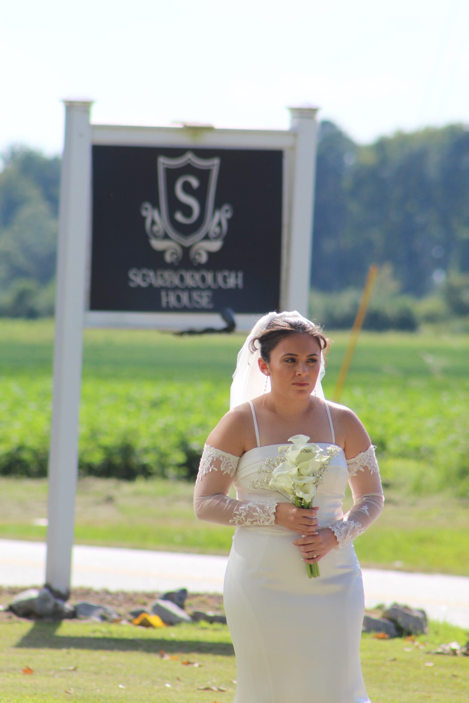 Bride in a white off-shoulder gown with lace gloves and veil holding a white calla lily bouquet in front of the Scarborough House estate sign surrounded by green fields.