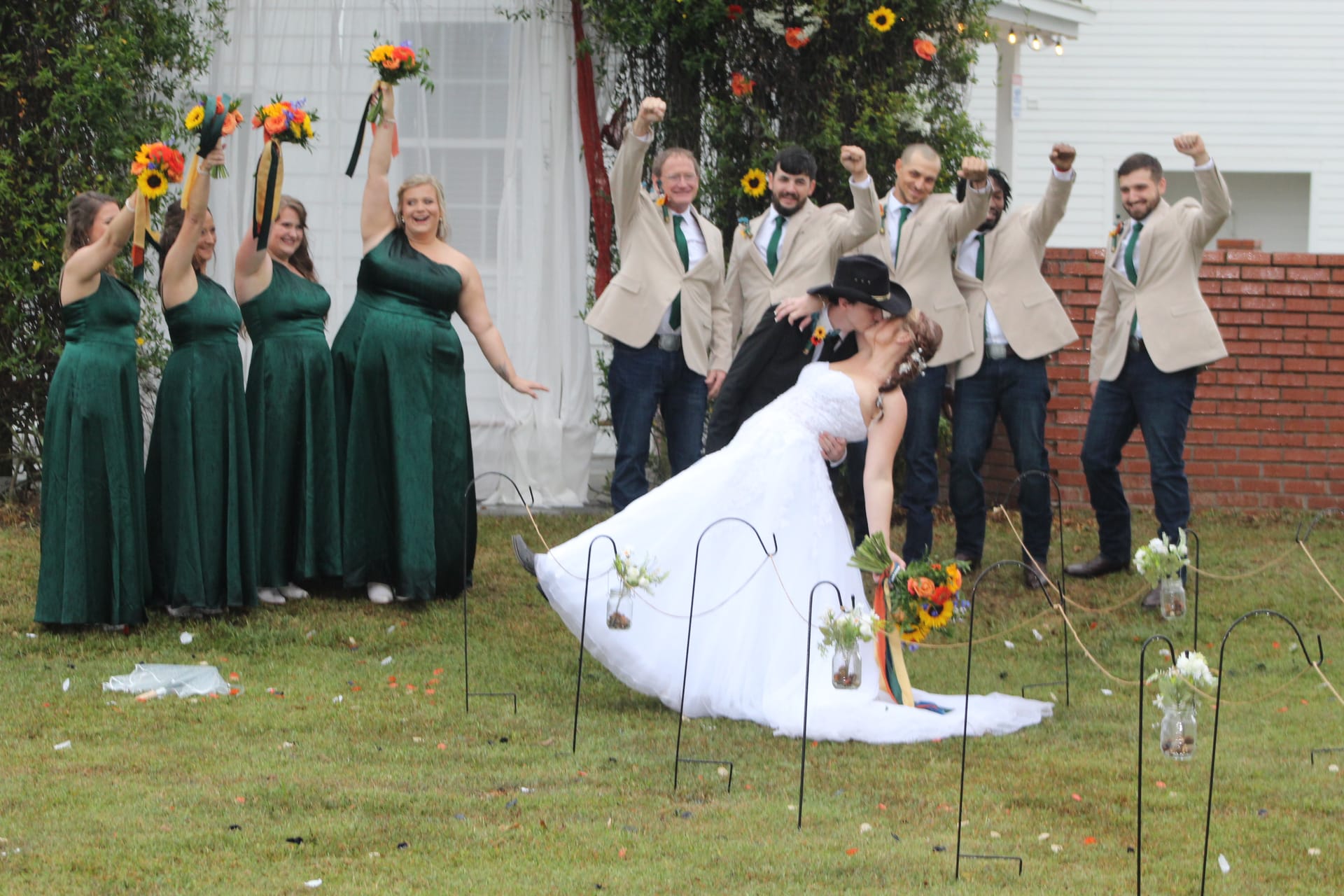 Groom dips bride in a kiss while their cheering wedding party in emerald green dresses and tan blazers celebrates behind them on the ceremony lawn at Scarborough House.
