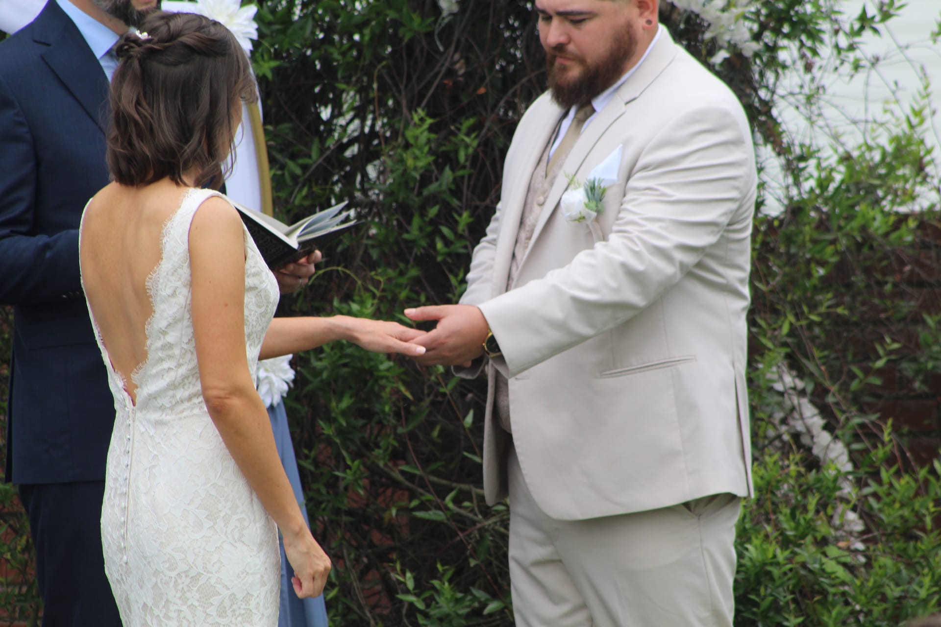 Groom in a tan suit gently places a ring on his brides hand during their outdoor wedding ceremony at Scarborough House, with lush greenery and white floral accents in the background.