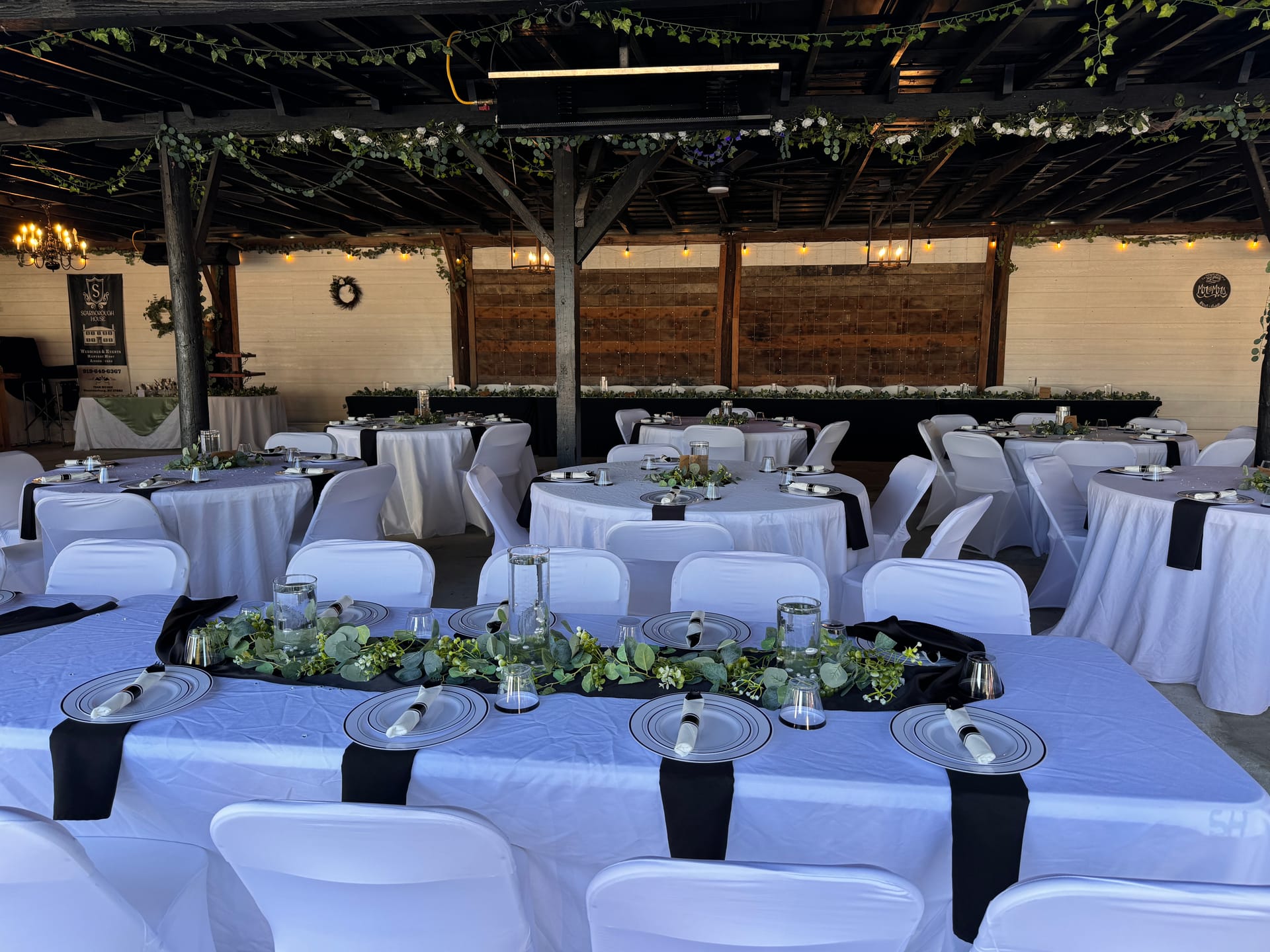 Elegantly decorated reception tables with white linens, black runners, greenery garland centerpieces, and silver charger plates inside a rustic pavilion adorned with string lights and trailing ivy at Scarborough House.