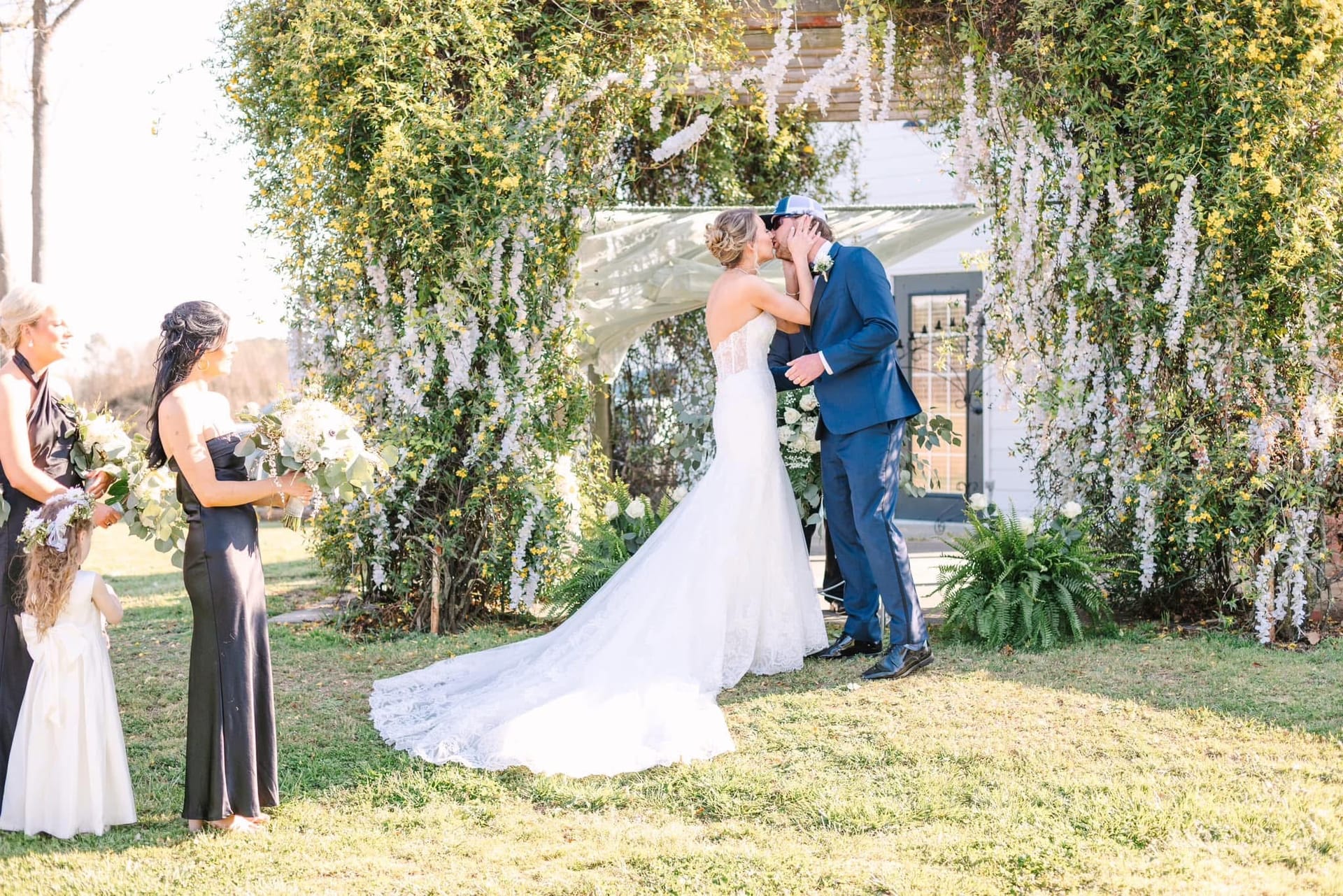 Bride and groom share their first kiss beneath a lush green floral arch on the ceremony lawn at Scarborough House, with bridesmaids in black gowns looking on.