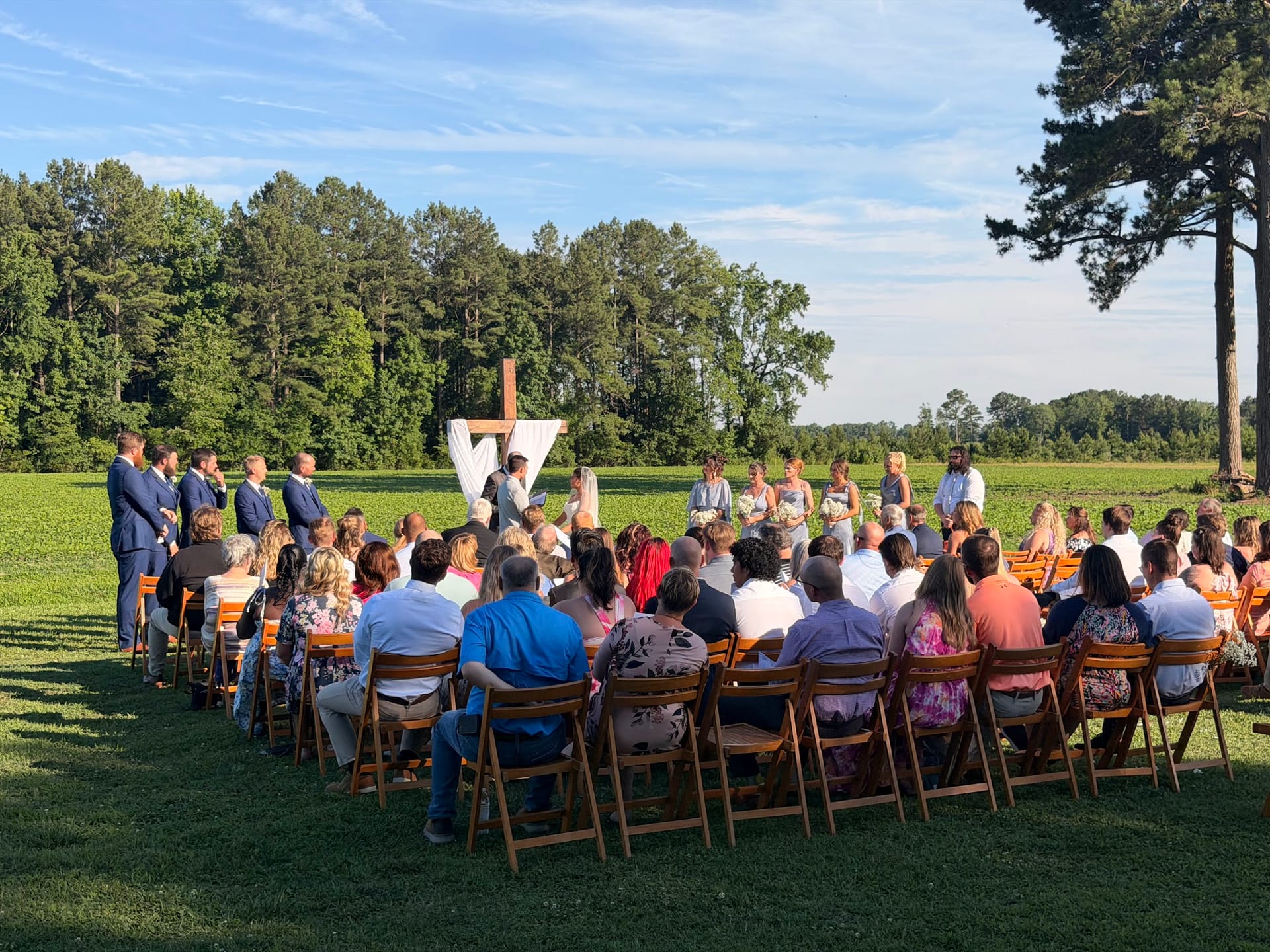 Outdoor wedding ceremony on a sunlit lawn at Scarborough House with guests seated in wooden chairs facing the couple at a rustic wooden cross altar surrounded by green fields and trees.
