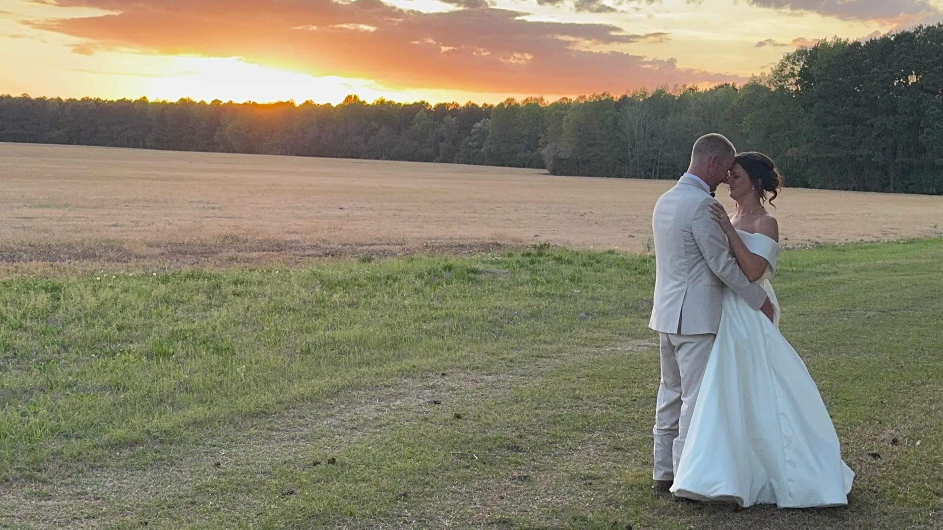 Bride in an off-the-shoulder white gown and groom in a light tan suit share an intimate embrace at golden hour overlooking Scarborough House's sweeping farmland fields beneath a vivid orange and gold sunset sky.