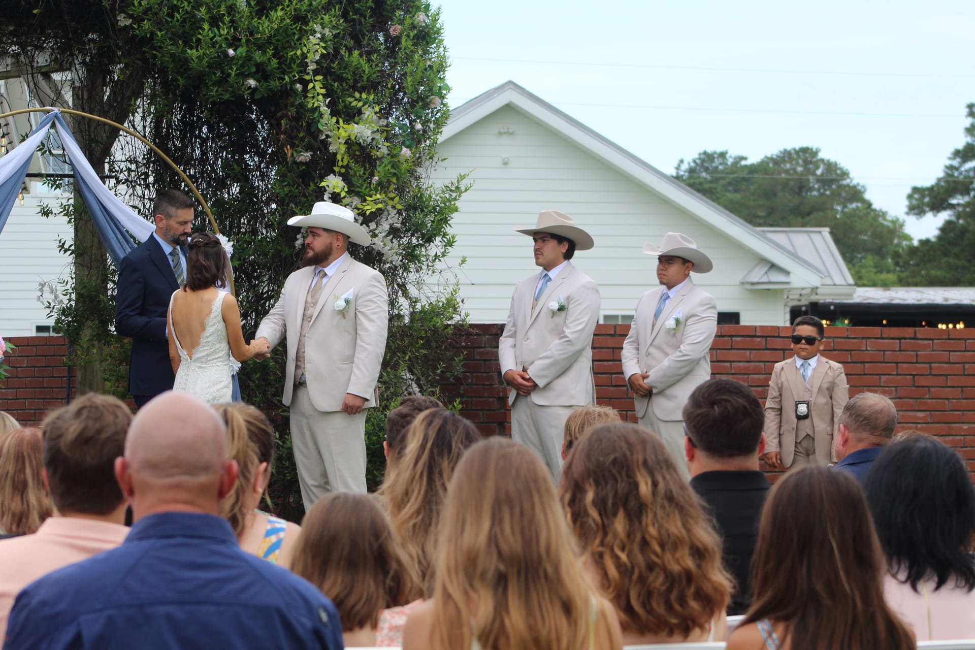 Three groomsmen in tan suits and cowboy hats stand at the outdoor altar during a western-themed wedding ceremony at Scarborough House, with a floral arch and white historic building in the background.