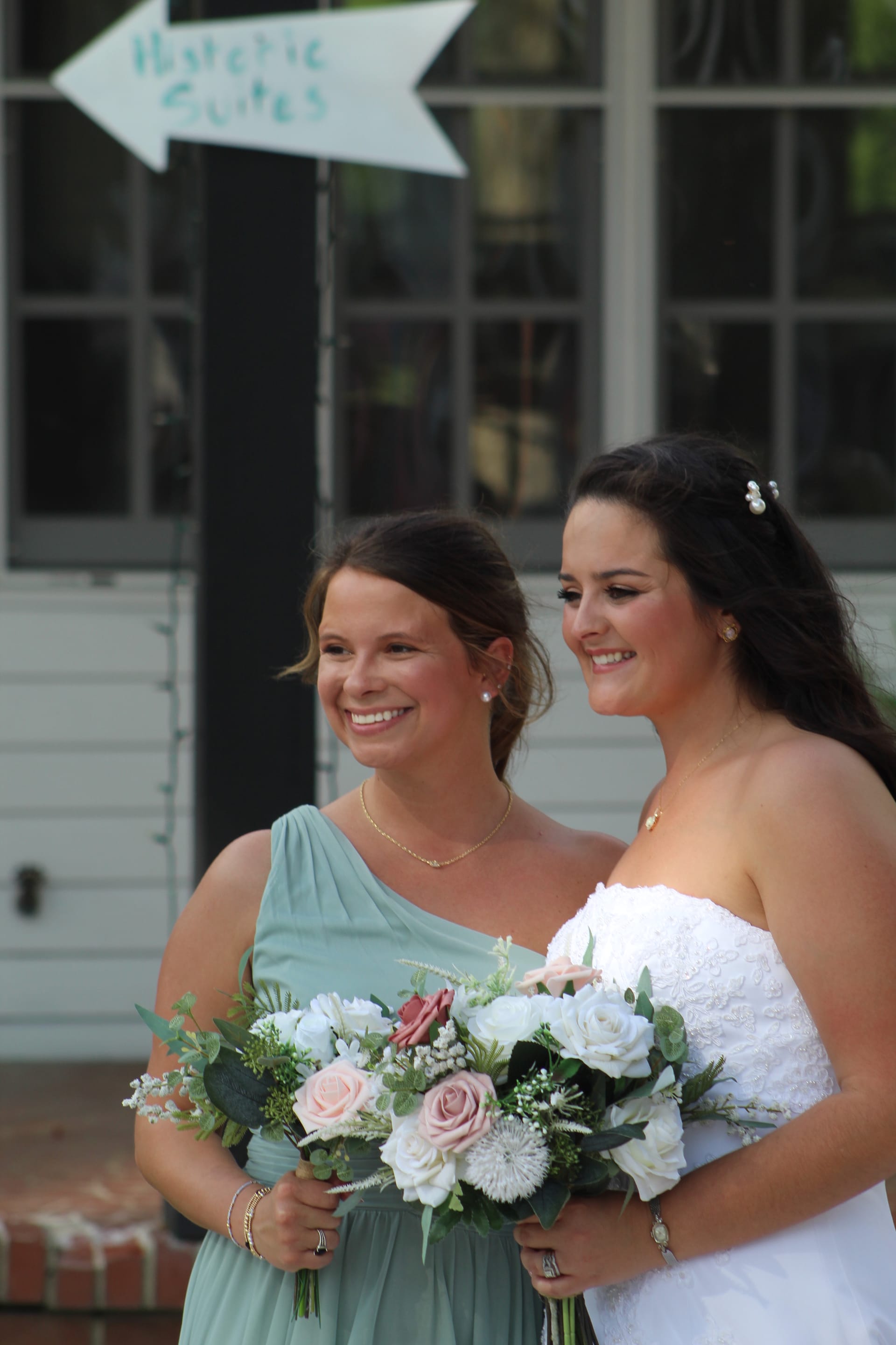 A smiling bride in a white strapless lace gown and her bridesmaid in a sage green one-shoulder dress hold pink and white floral bouquets outside a historic estate venue.