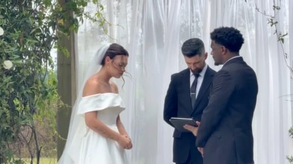 Bride in an off-shoulder white gown and groom stand before an officiant reading vows beneath flowing white drapery and greenery at a garden ceremony.