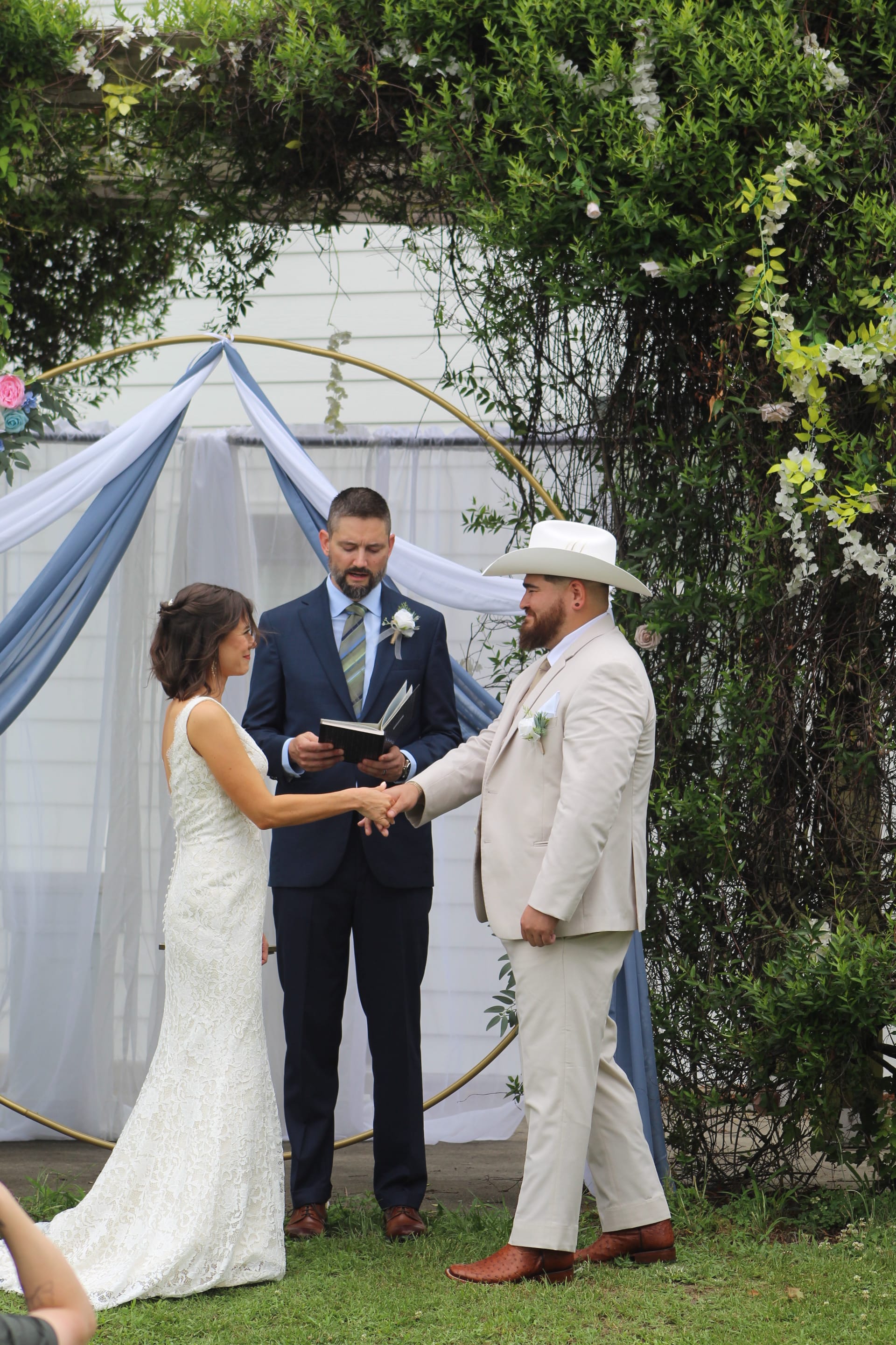Bride and groom hold hands during outdoor wedding vows beneath a gold circular arch draped in blue and white fabric with lush greenery at Scarborough House.