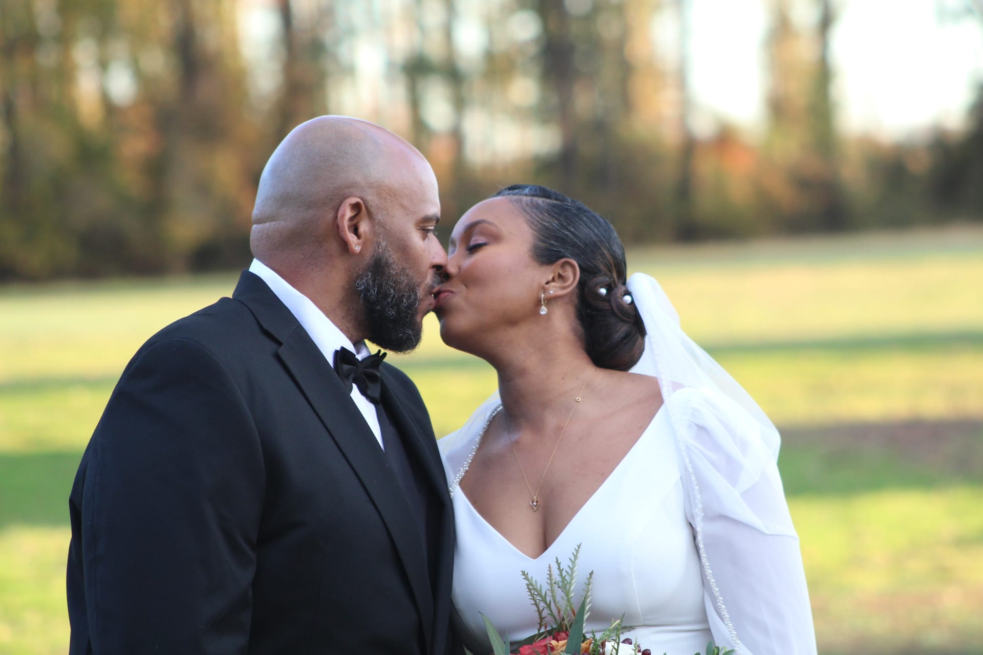 Bride in white gown and veil shares a tender kiss with groom in black tuxedo on the sunlit ceremony lawn at Scarborough House wedding venue.