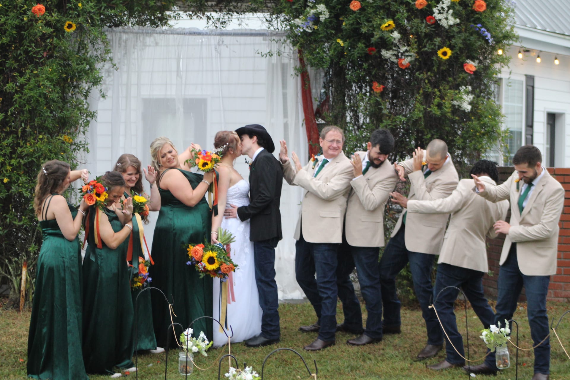 Bride and groom share their first kiss beneath a floral-adorned garden arch as bridesmaids in emerald gowns and groomsmen in cream blazers cheer and applaud on the ceremony lawn at Scarborough House.