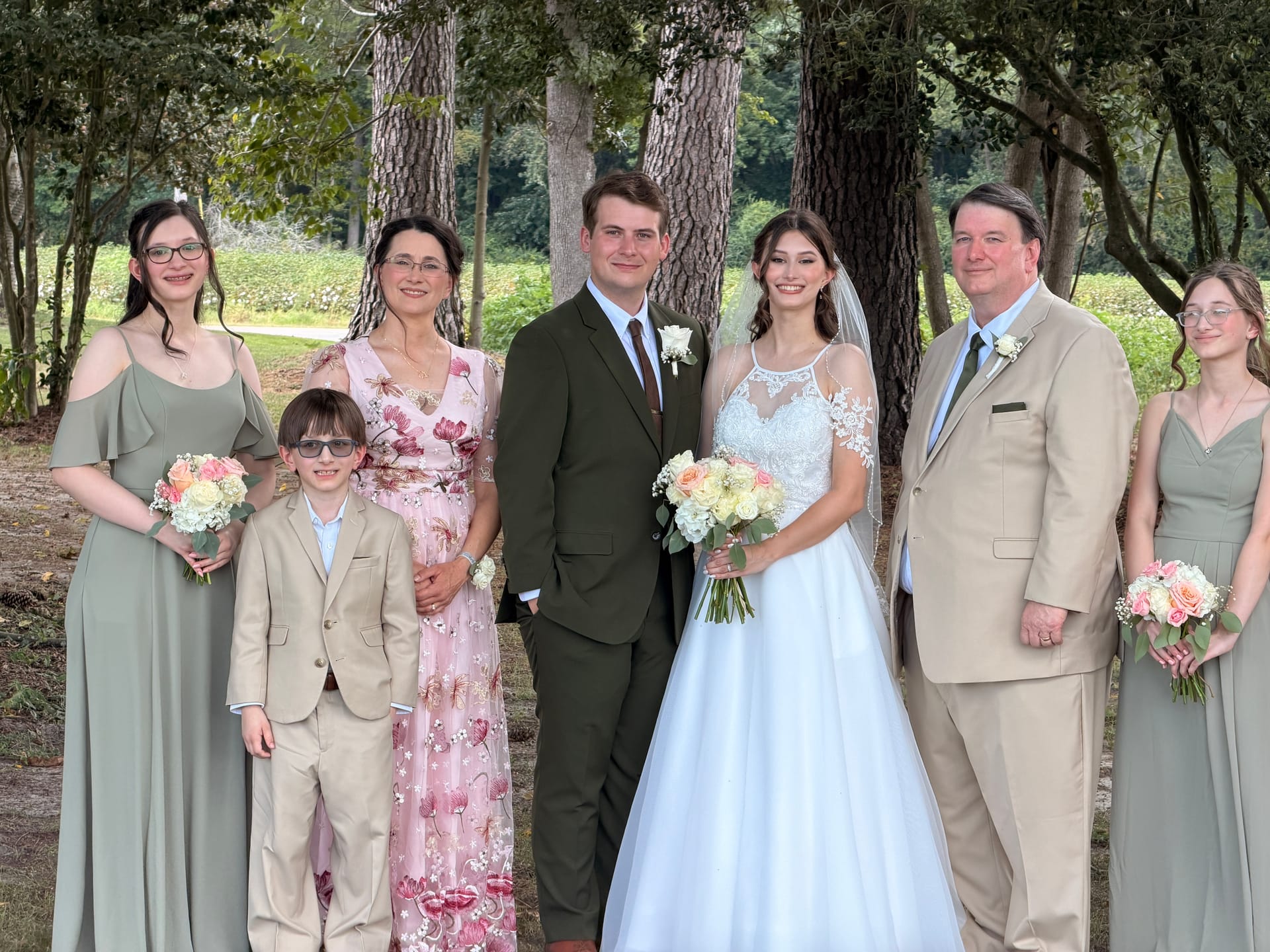 Bride in white lace gown and groom in olive suit pose with family members in sage bridesmaid dresses and tan suits beneath tall oak trees at Scarborough House wedding venue.