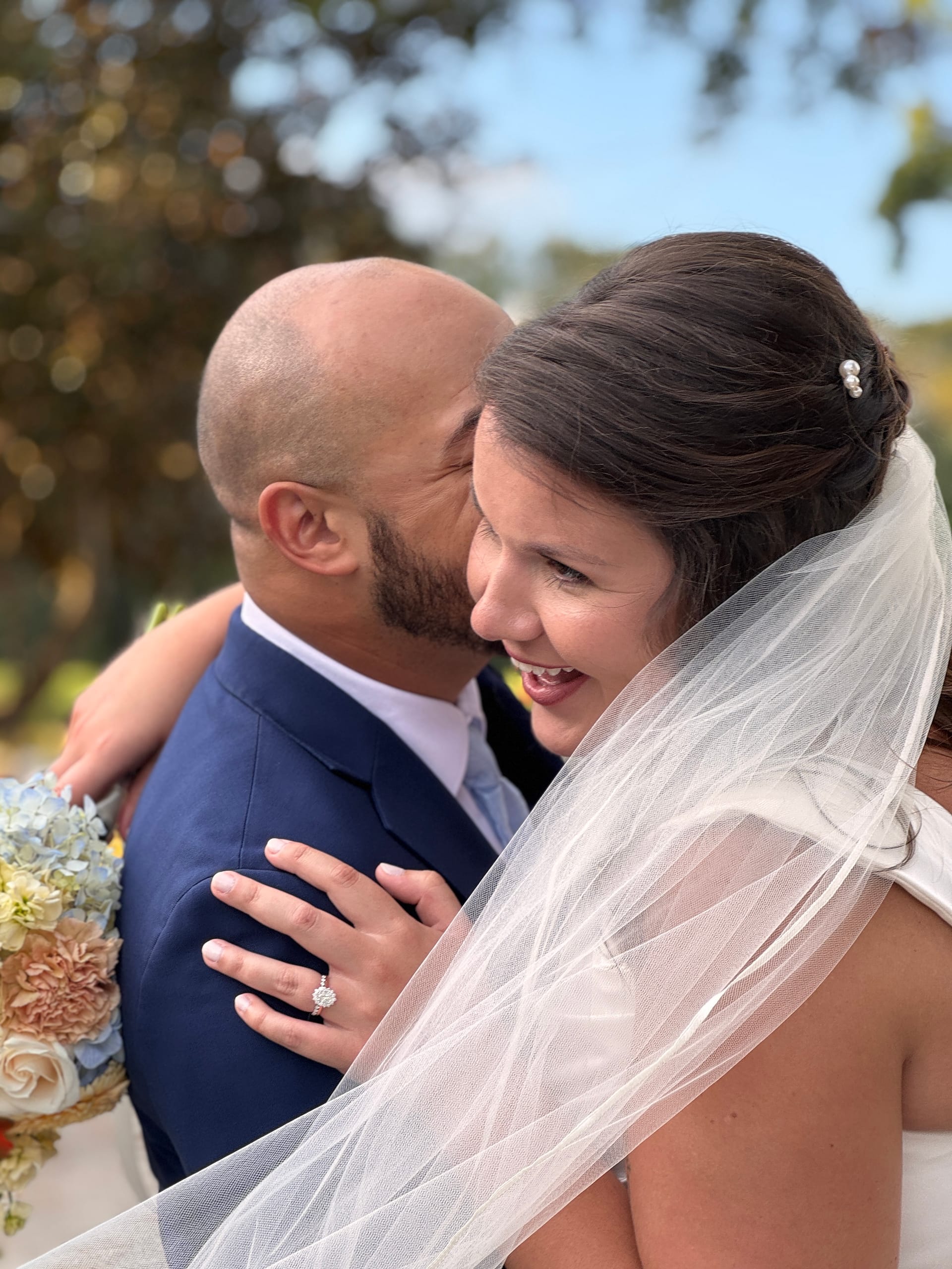 Smiling bride in a white veil laughs as her groom in a navy suit kisses her cheek beneath oak trees at Scarborough House, her diamond engagement ring catching the light against his shoulder.