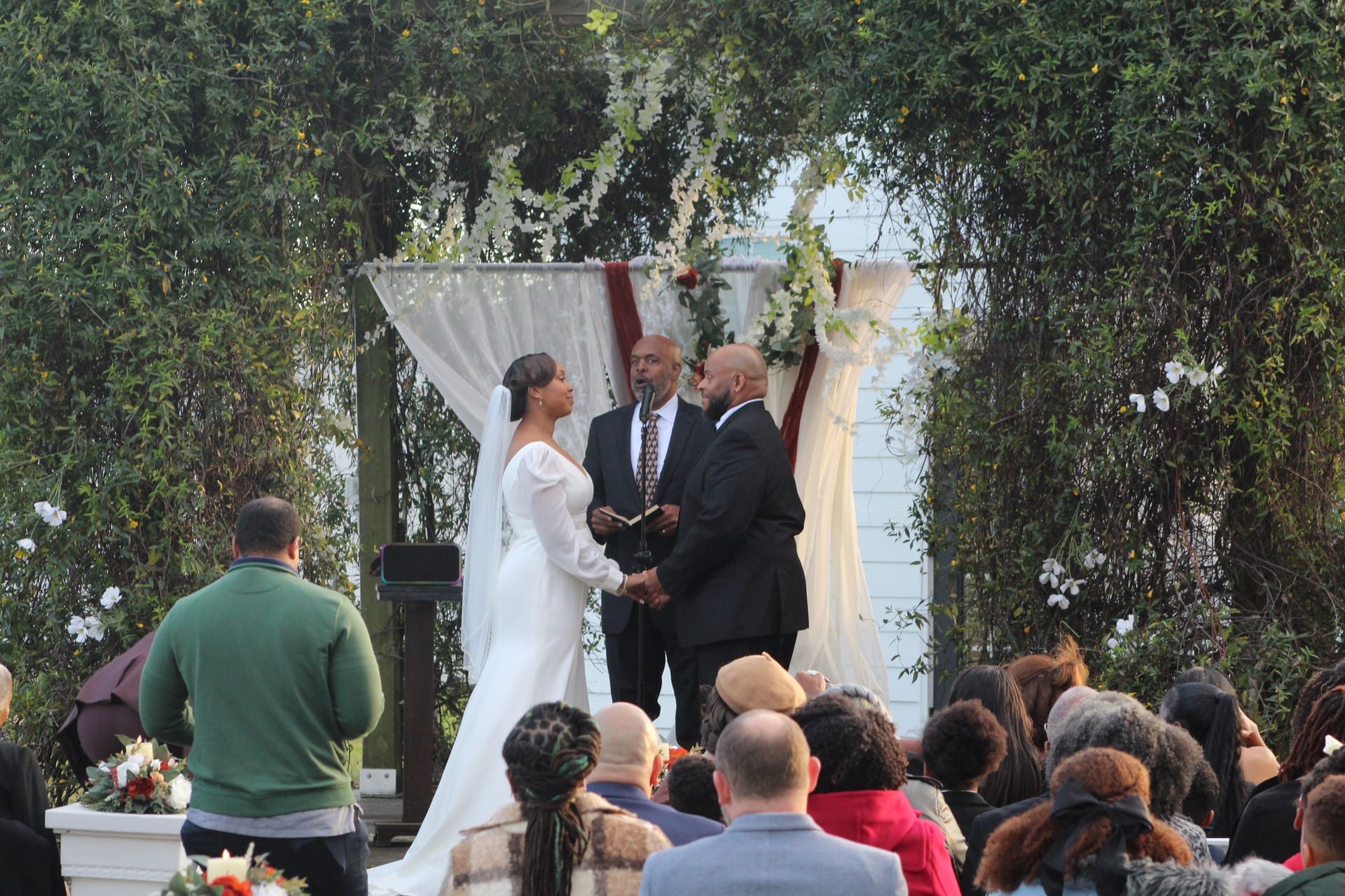 Bride in a white long-sleeve gown and groom in a black suit exchange vows beneath a draped floral arch surrounded by lush greenery at Scarborough House ceremony lawn.