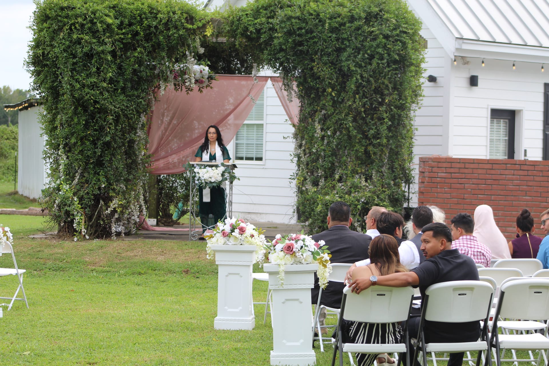 Outdoor wedding ceremony at Scarborough House with officiant speaking at a lush greenery arch adorned with blush draping and florals while guests are seated on white chairs flanked by white pedestal floral arrangements.