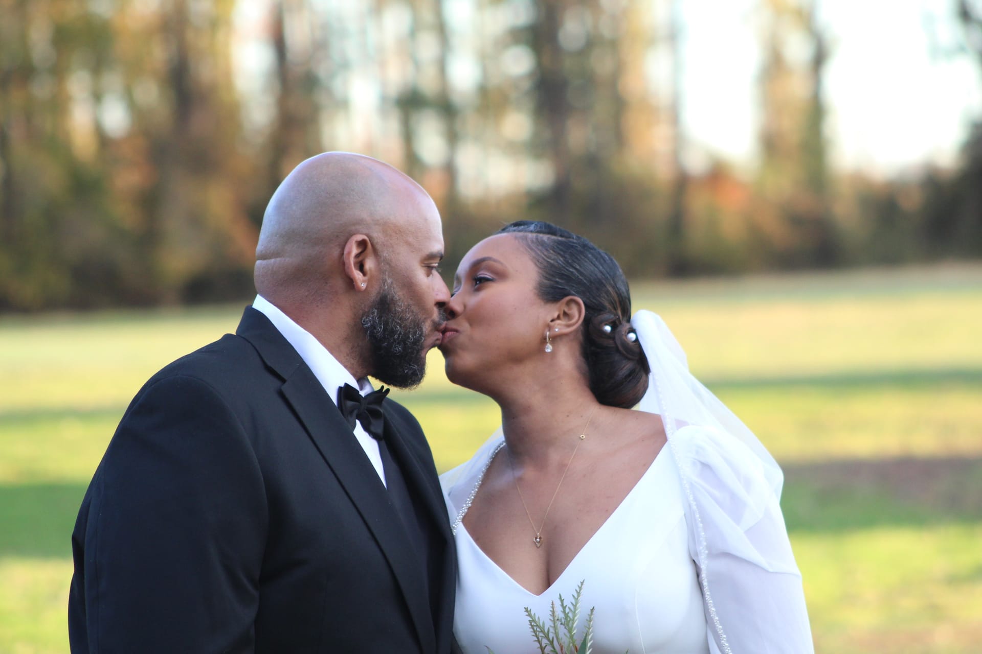 Bride in a white gown and veil and groom in a black tuxedo share a romantic kiss during outdoor portrait session on a sunlit field surrounded by autumn trees.
