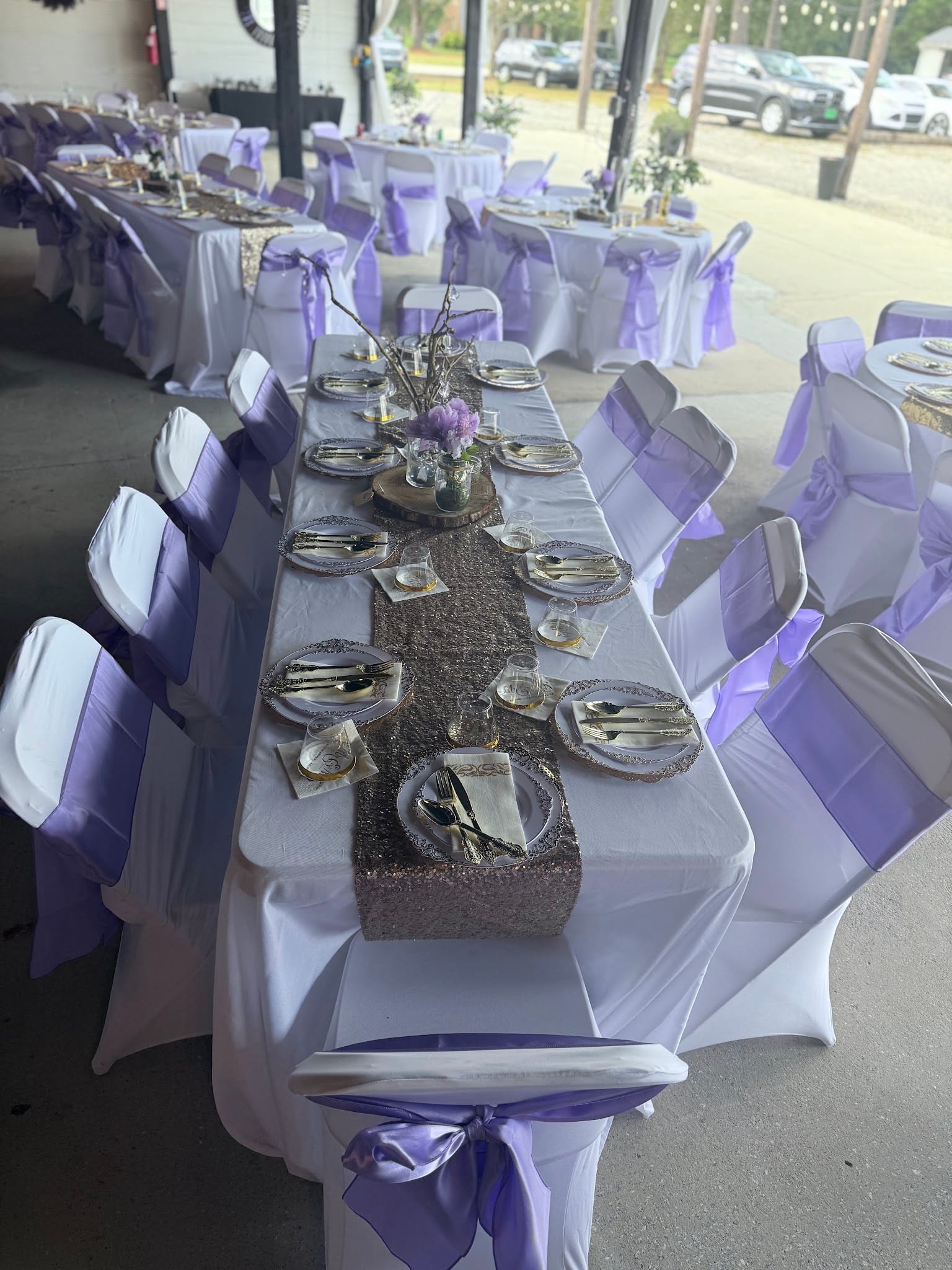 Elegantly set rectangular reception table with white linens, a glittery rose gold table runner, gold-rimmed charger plates, lavender floral centerpieces, and white chair covers tied with purple sashes at Scarborough House wedding venue.
