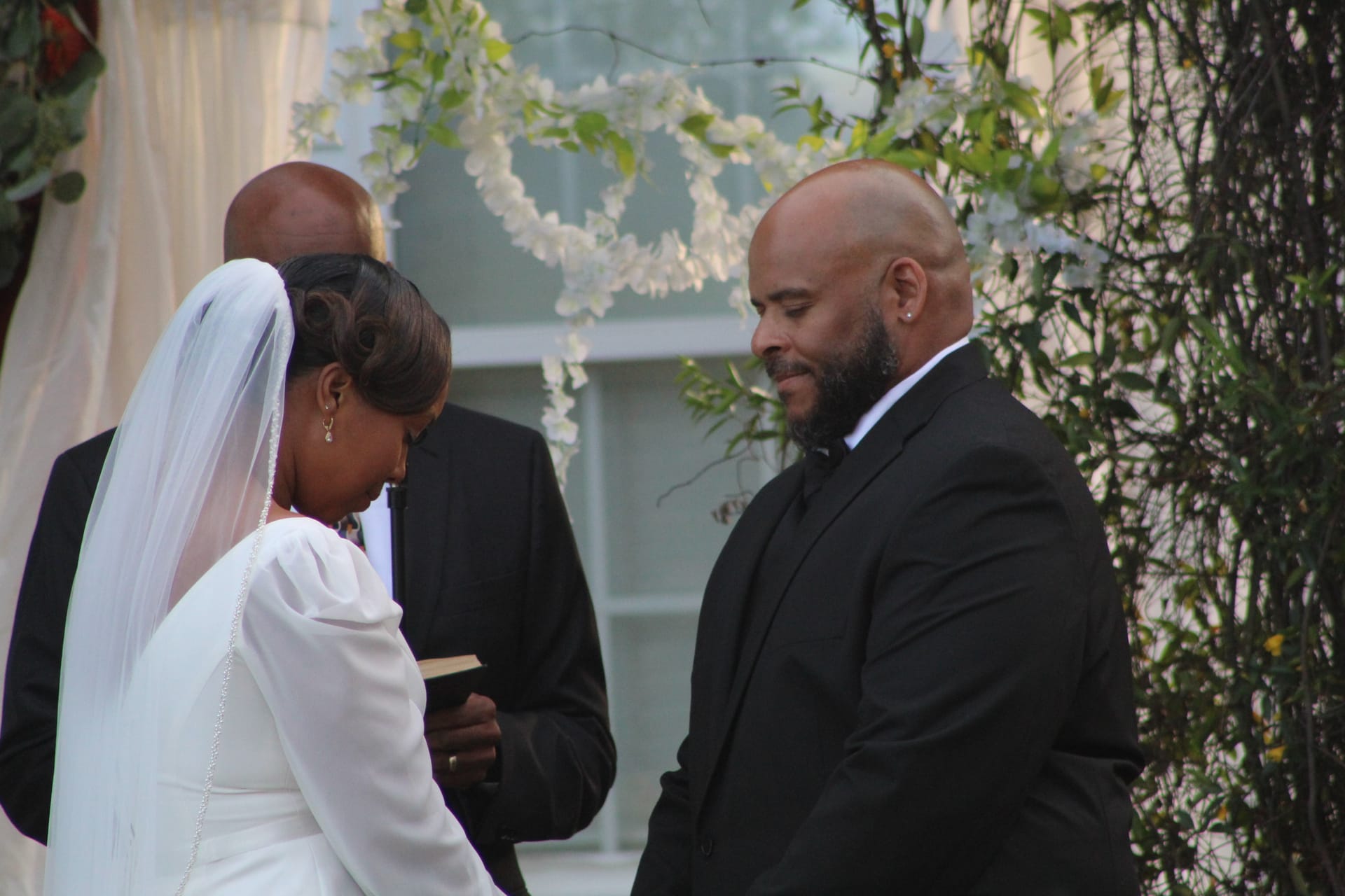 Bride in white gown and veil reads vows from a book as her groom in a black suit looks on with a warm smile beneath a floral heart arch at Scarborough House.