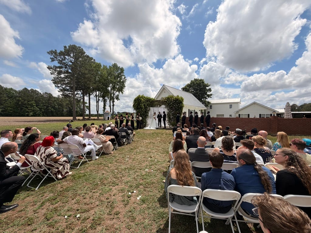 Guests seated in white folding chairs on the ceremony lawn at Scarborough House watch a couple exchange vows beneath a lush floral arch with the white chapel and farmland in the background under a partly cloudy summer sky.