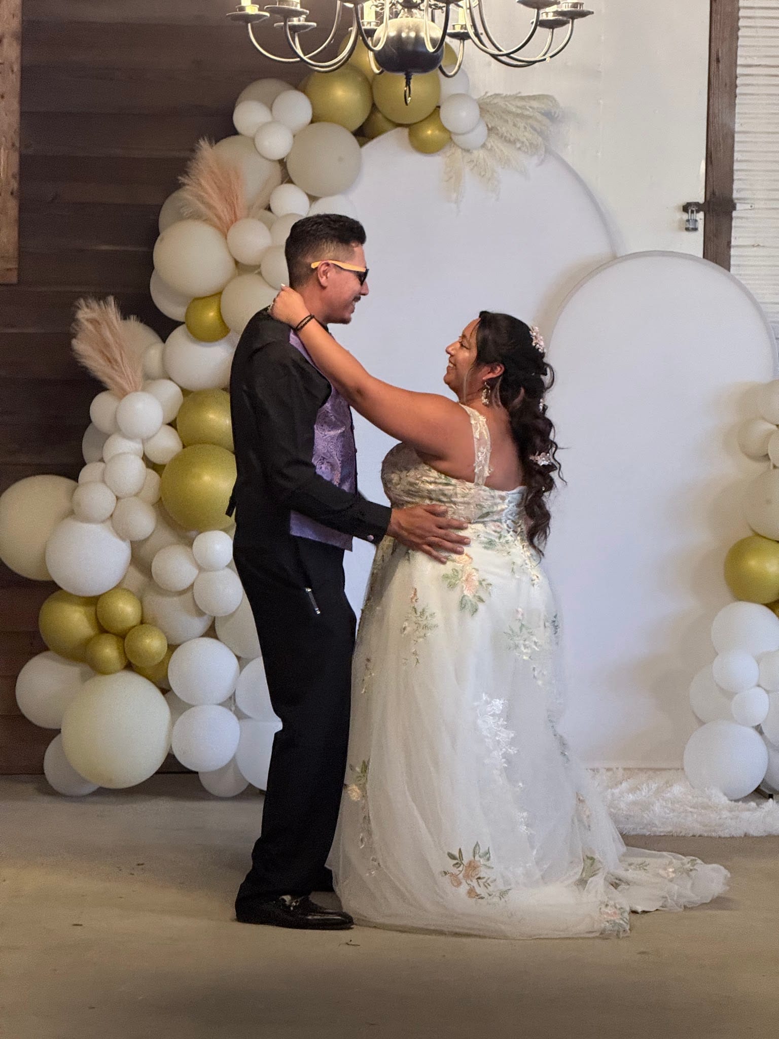 Bride in a floral embroidered gown and groom in black suit share a joyful first dance in front of a white arch adorned with gold and ivory balloon garlands and pampas grass.