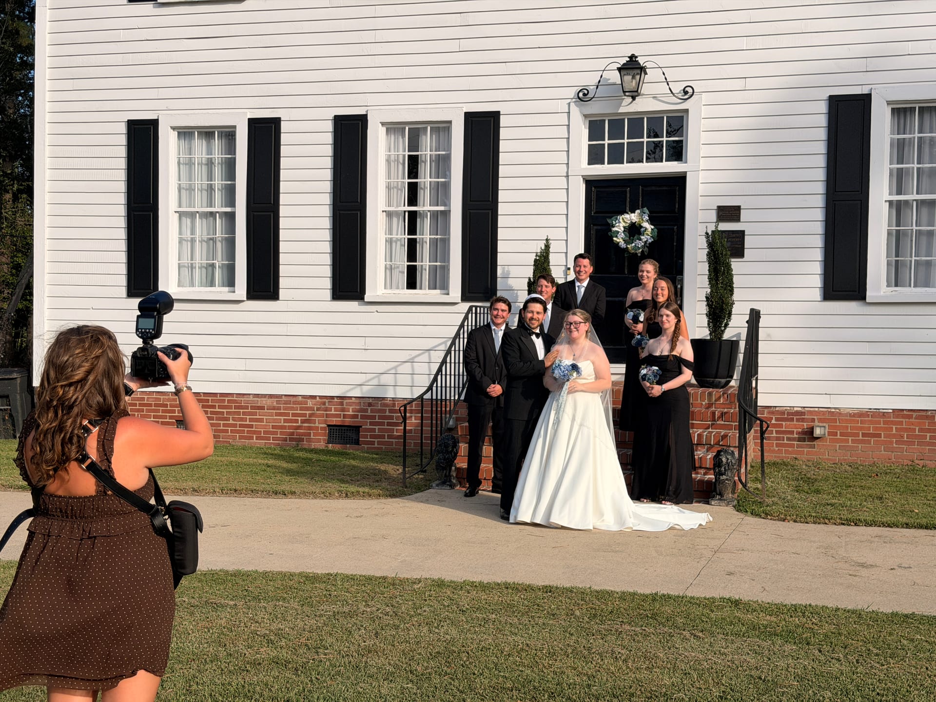 A wedding party including the bride in a white gown and bridesmaids in black dresses poses on the front steps of the historic 1821 Scarborough House manor while a photographer captures the moment.