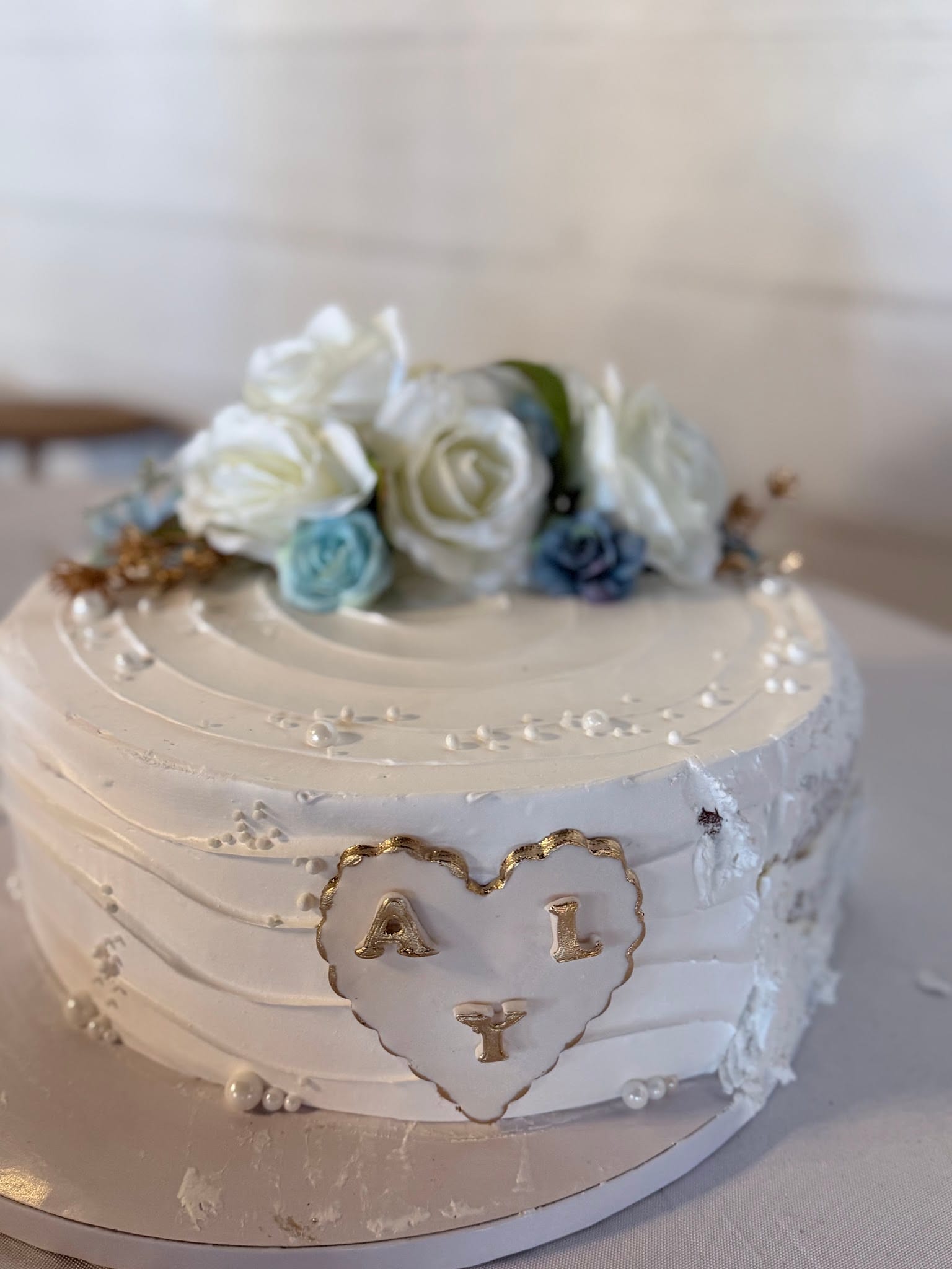 Close-up of an elegant white buttercream wedding cake adorned with white and dusty blue roses and a gold-edged heart monogram reading ALY at Scarborough House.