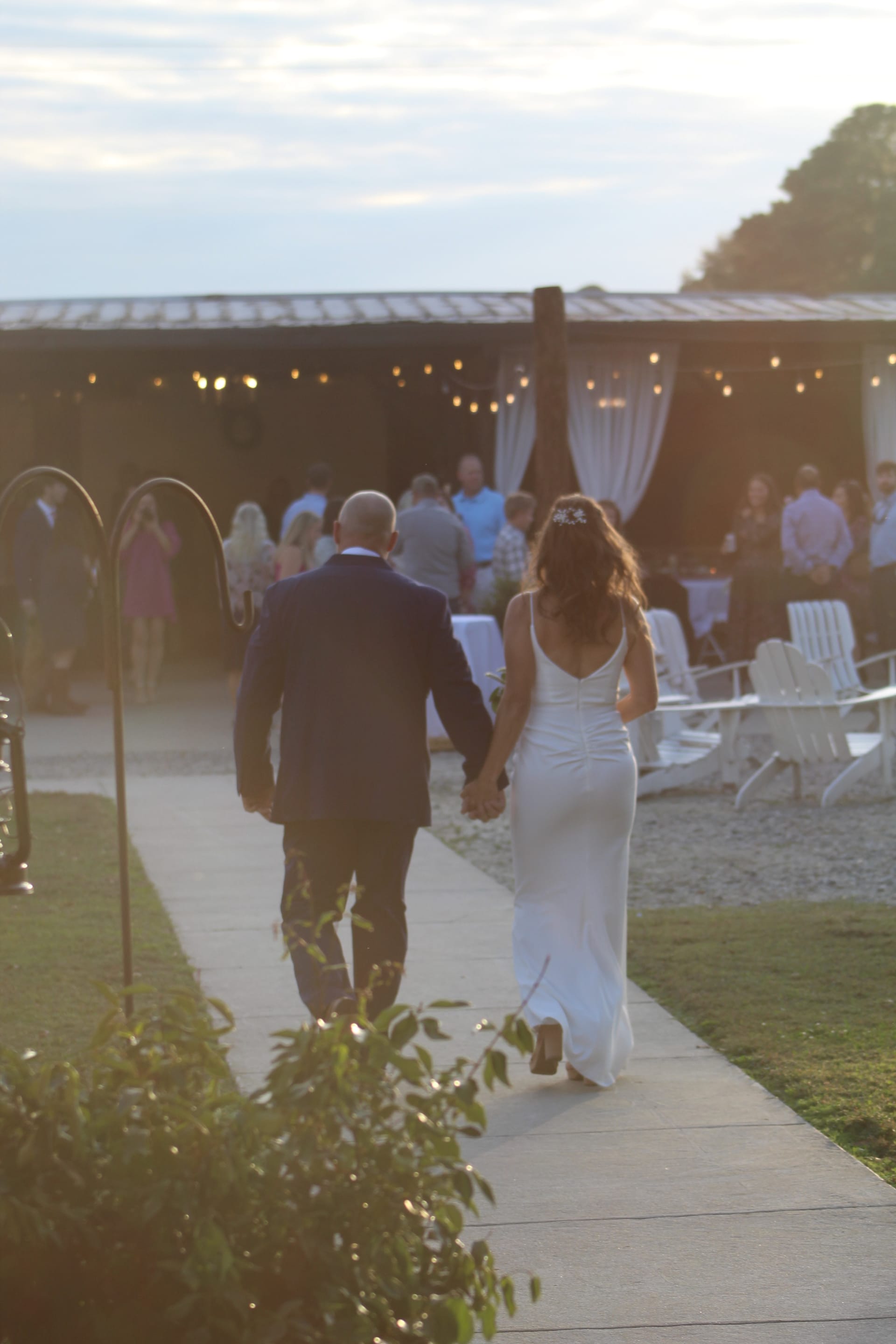 Bride in a fitted white gown and groom in a dark suit walk hand in hand along a garden path toward a string-light-draped pavilion filled with wedding guests at golden hour.