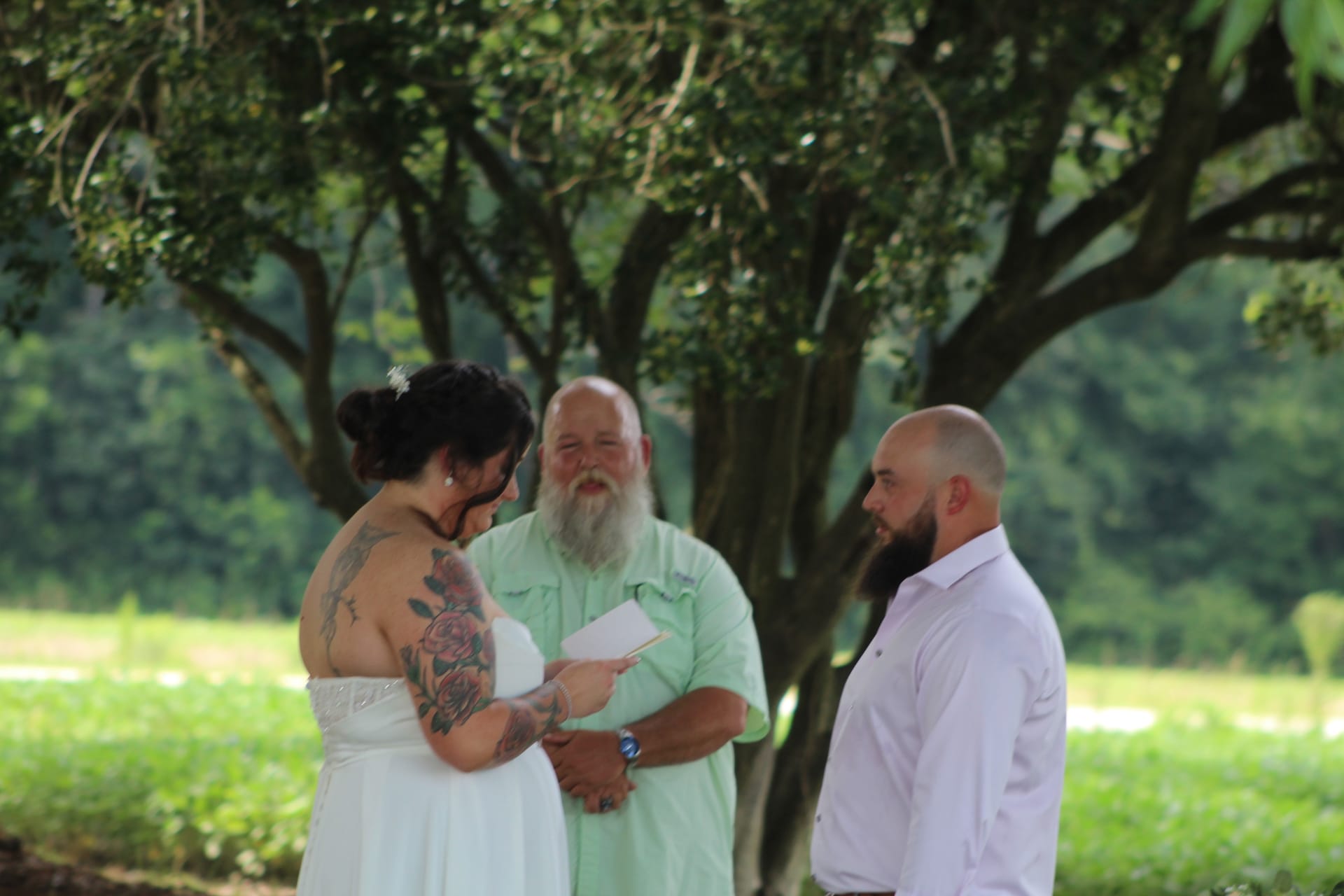 A tattooed bride in a white strapless gown reads her vows to a bearded groom in a white shirt while an officiant with a long gray beard presides under towering oak trees at a lush outdoor estate wedding.