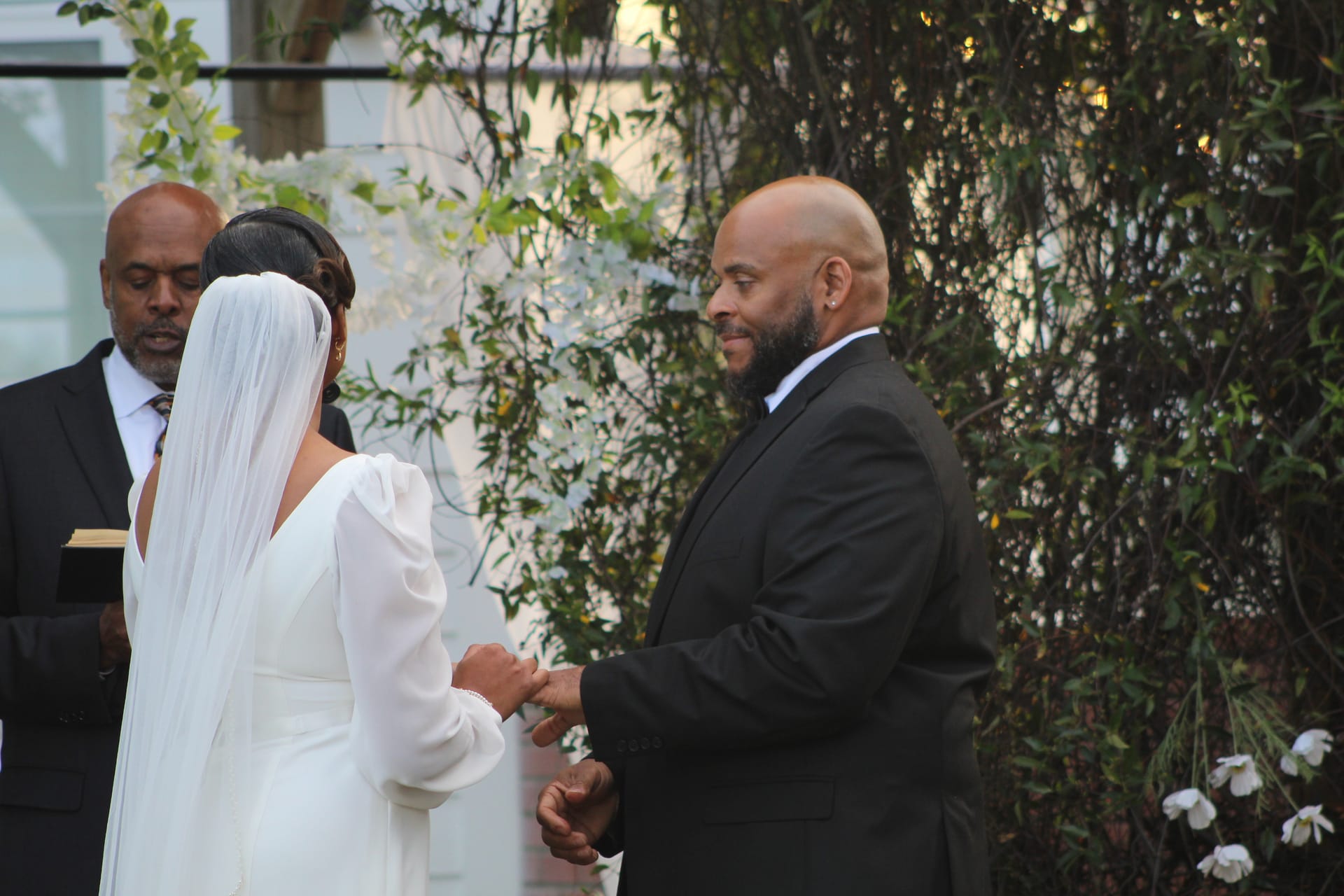 Groom smiles warmly while holding his bride's hands during outdoor wedding vows at a historic estate ceremony surrounded by lush greenery and a white floral arch.