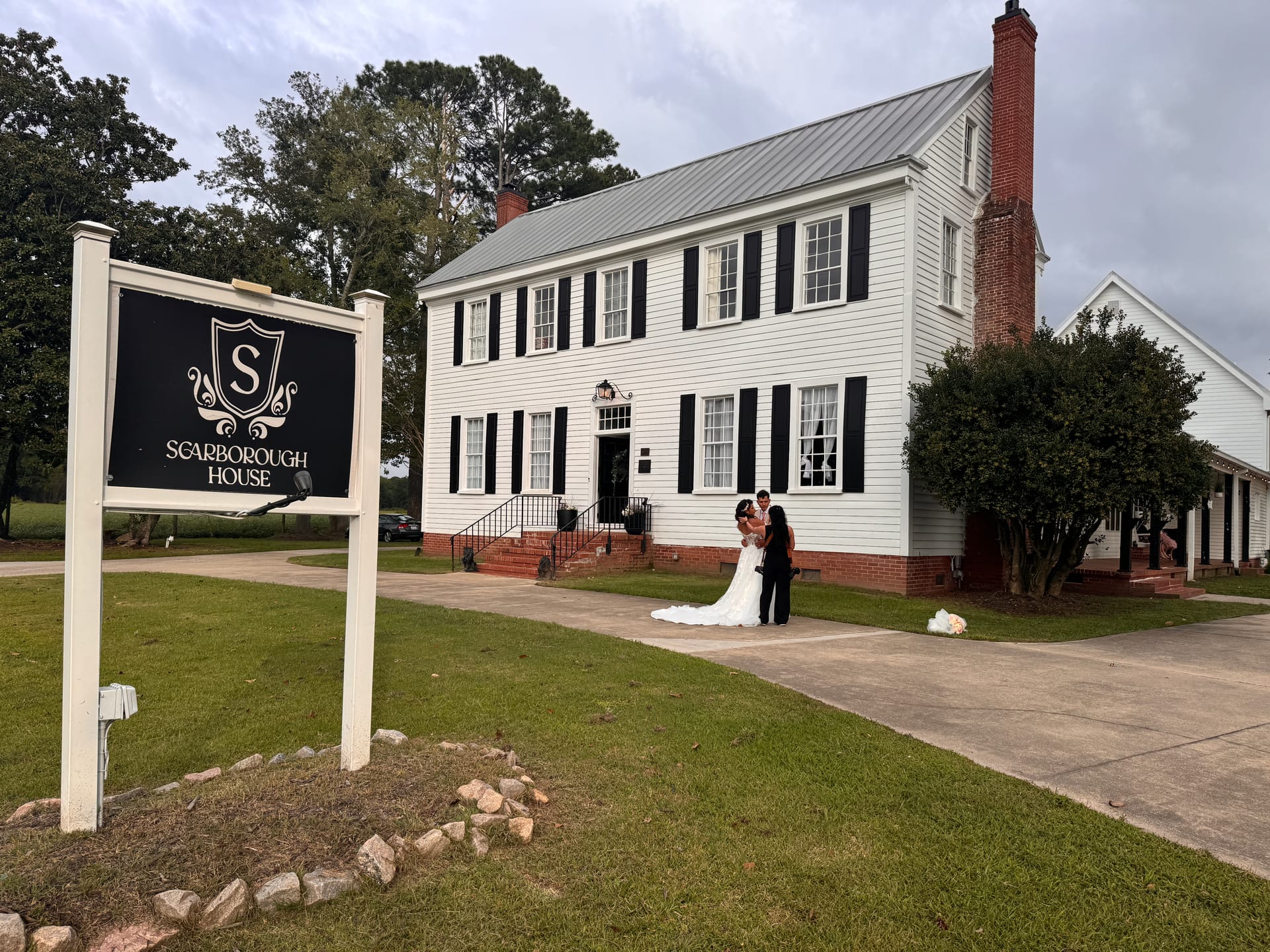Bride and groom embrace in front of the white historic Scarborough House manor with black shutters and brick chimney, framed by the venue entrance sign.