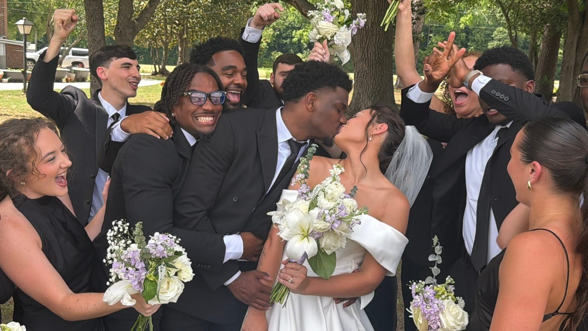 Bride and groom share a kiss while their jubilant wedding party cheers and celebrates around them beneath the oak trees at Scarborough House.