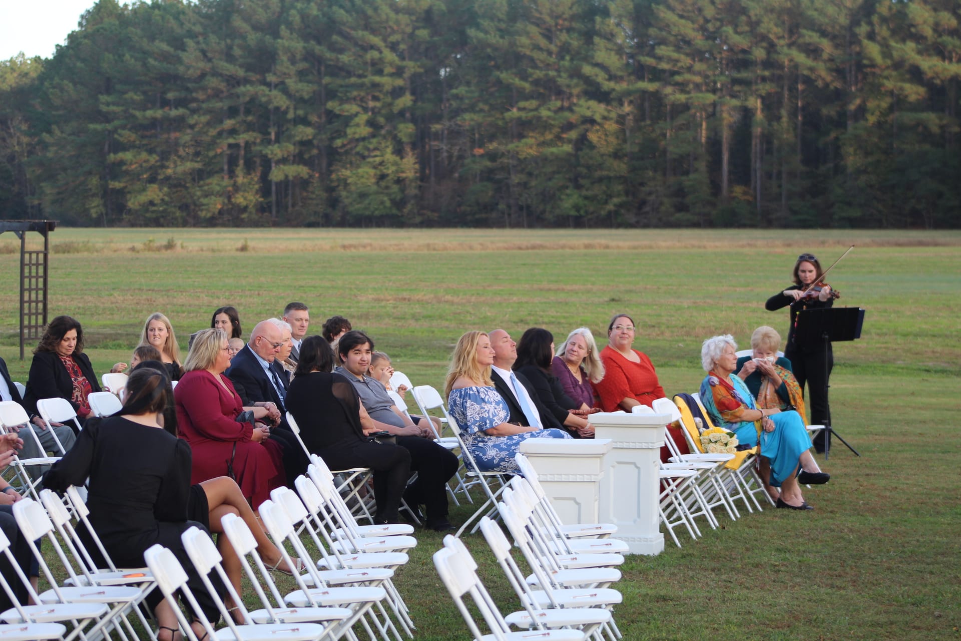 Wedding guests seated in white folding chairs on the open ceremony lawn at Scarborough House while a violinist performs at golden hour with pine forest in the background.
